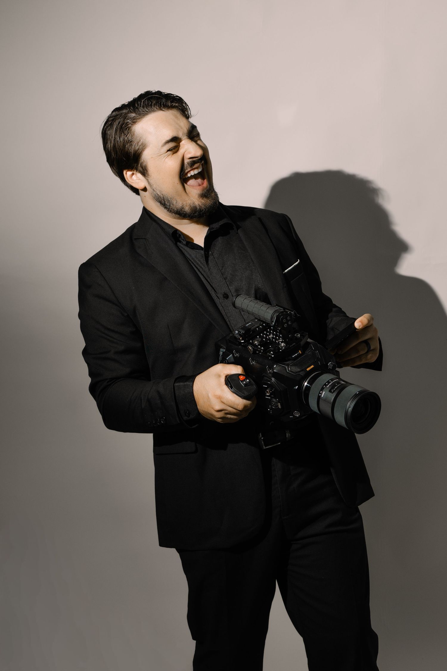A smiling man in a black suit holds a professional video camera in a studio setting.