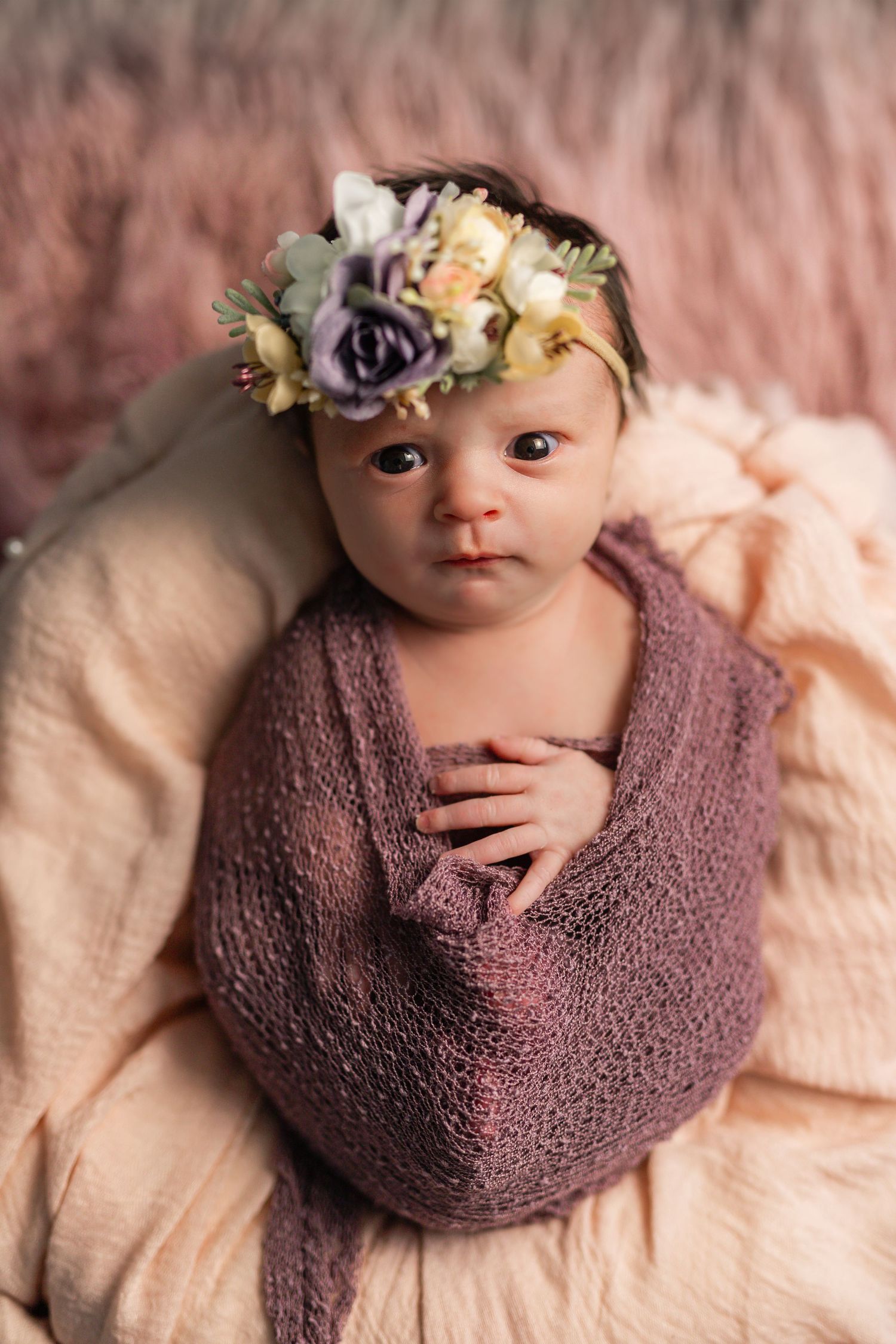 Newborn baby wrapped in purple knit swaddle wears a floral crown on a pink blanket.