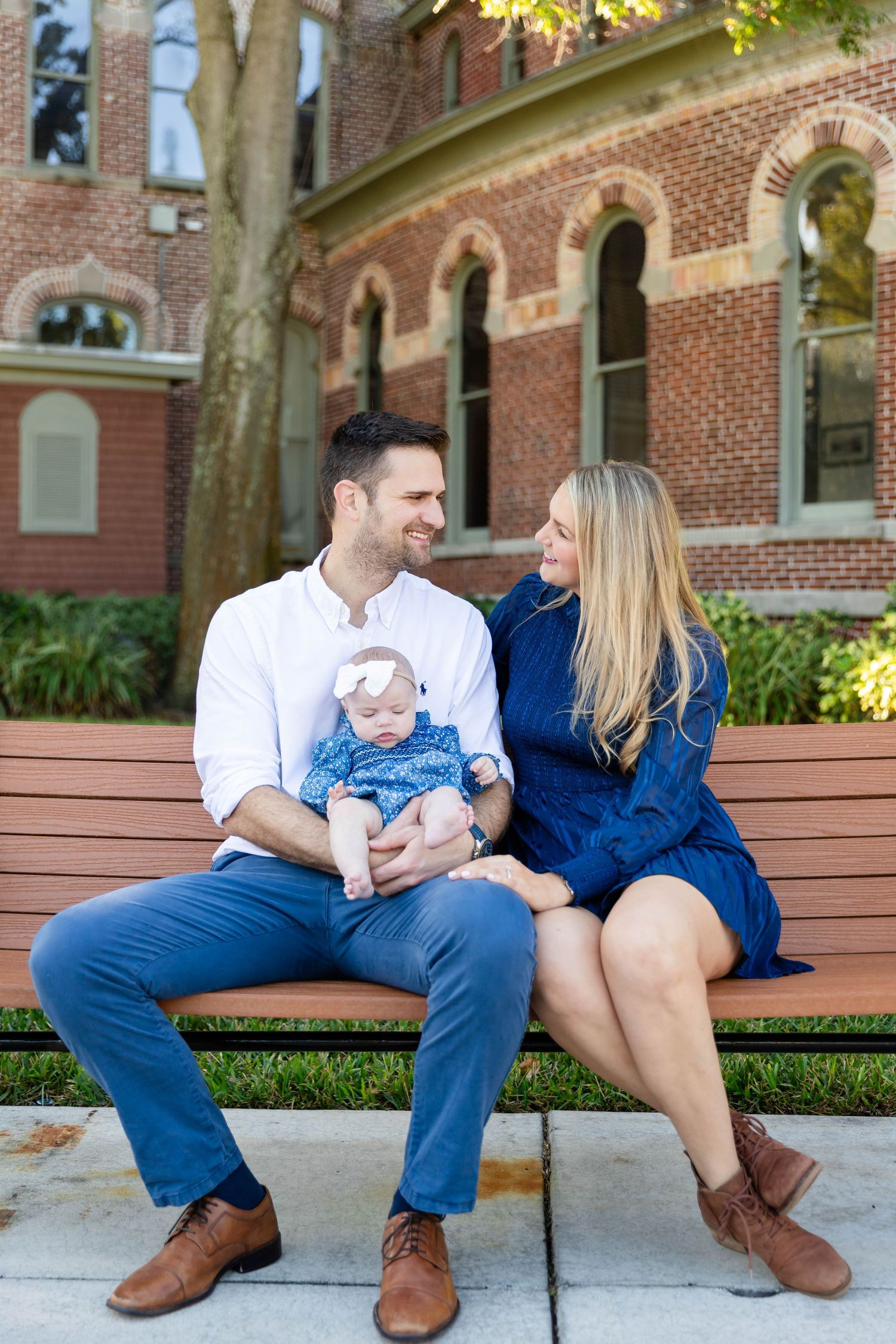 Young family sits on outdoor bench, father holding infant baby while mother looks on lovingly at the University of Tampa.