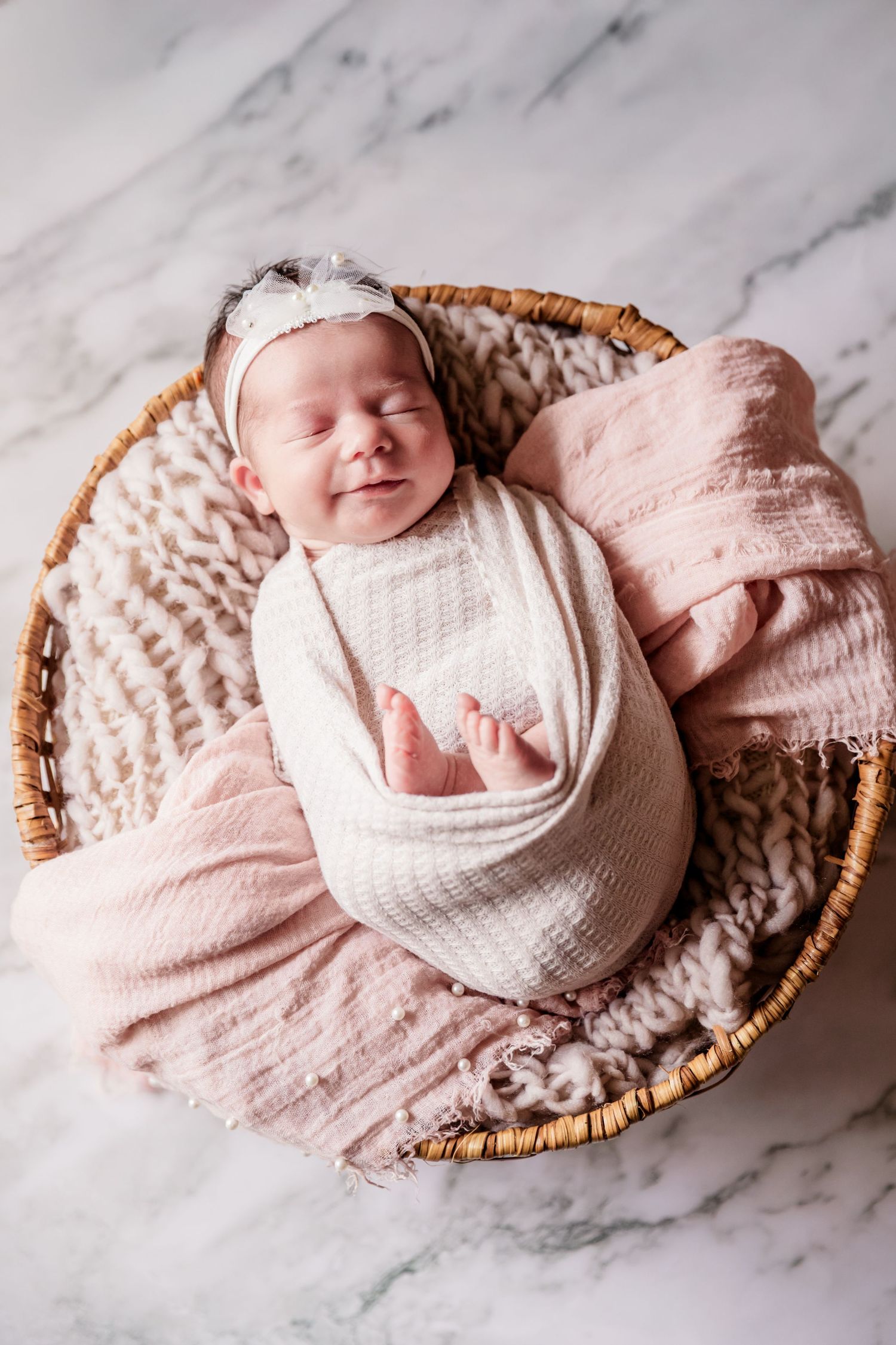 Peaceful newborn baby swaddled in white wrap resting in a wicker basket with pink blanket.