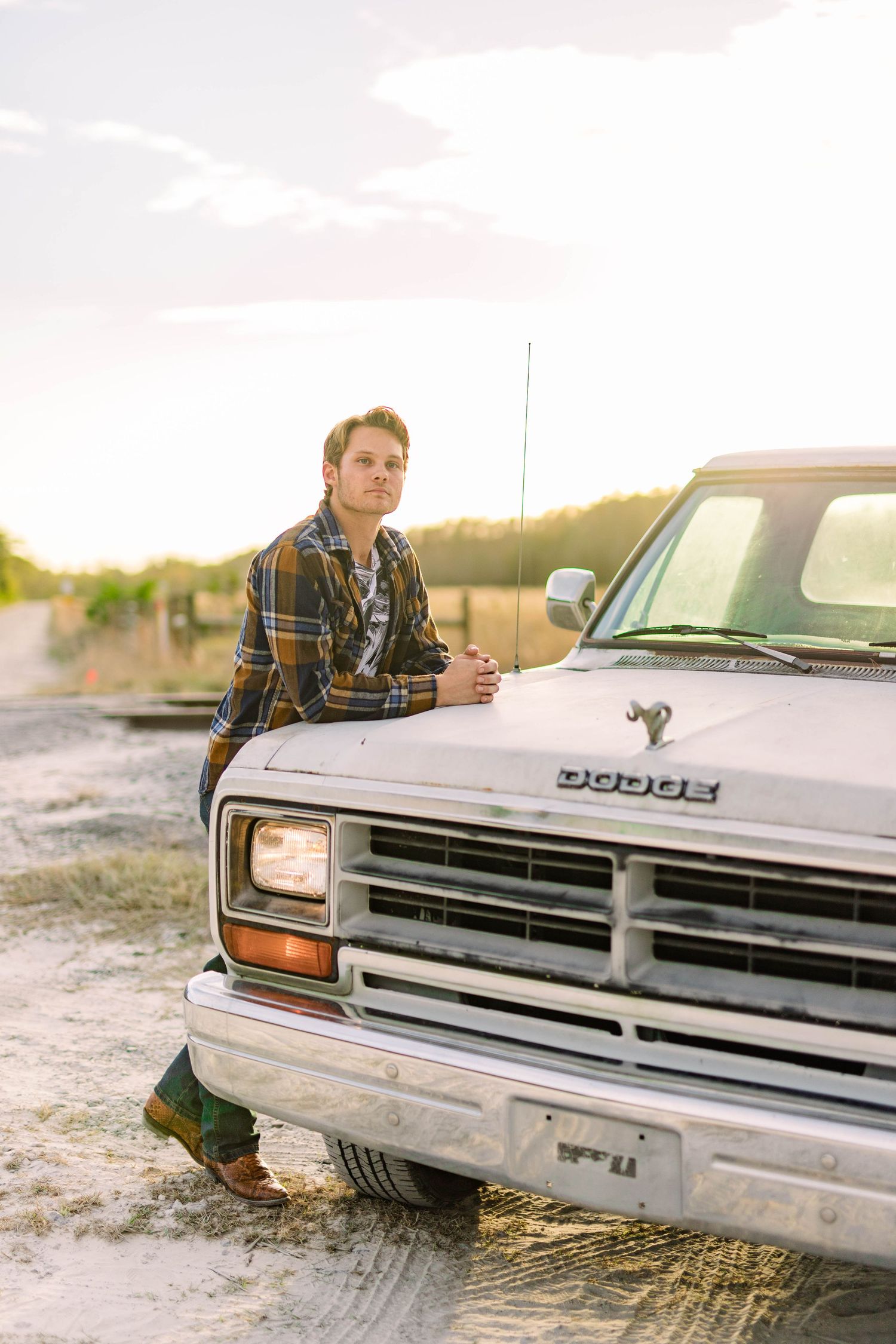 Young man in plaid shirt leaning against vintage white Dodge truck at golden sunset.
