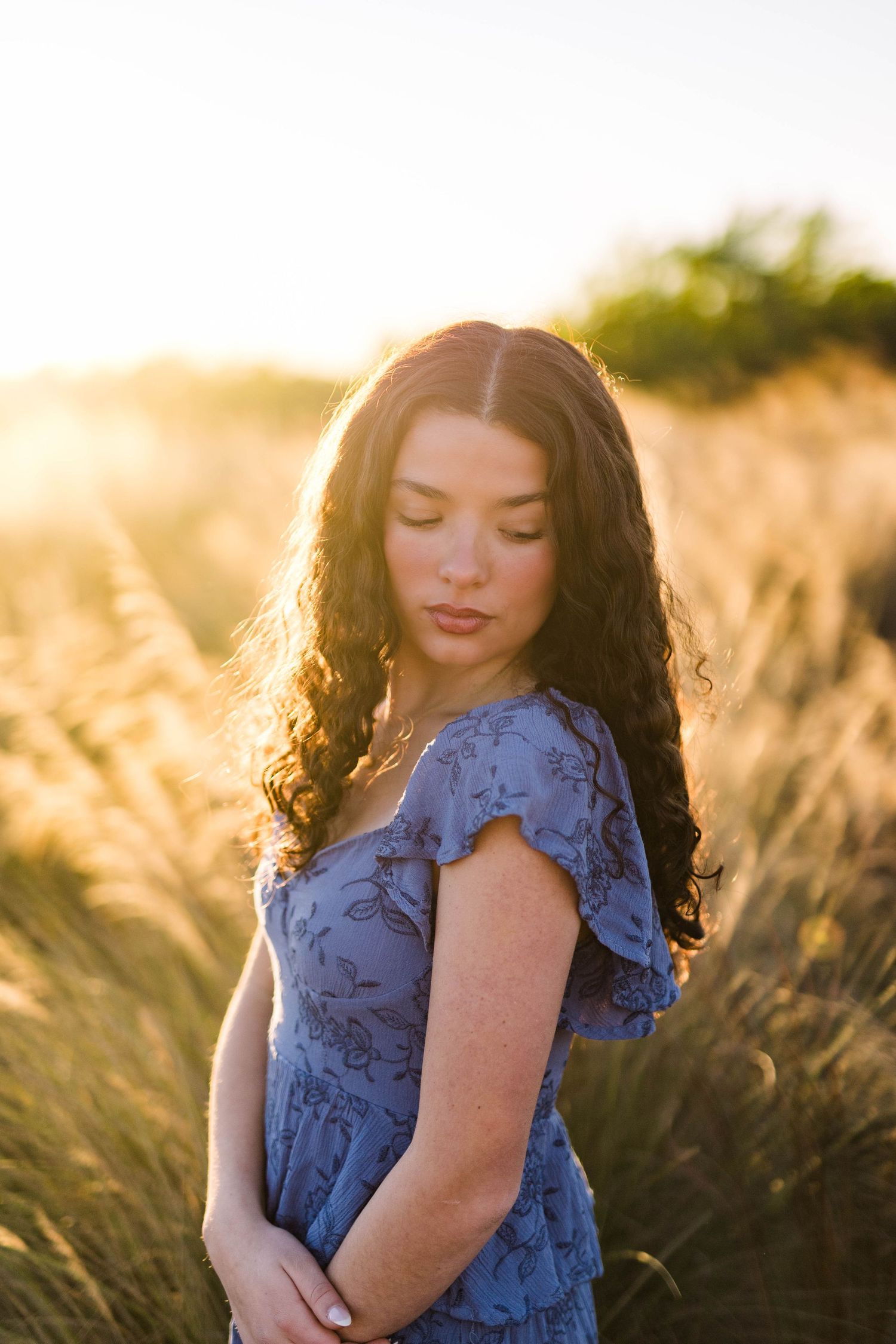 Young woman with curly dark hair in a blue floral dress standing in a golden wheat field at sunset.