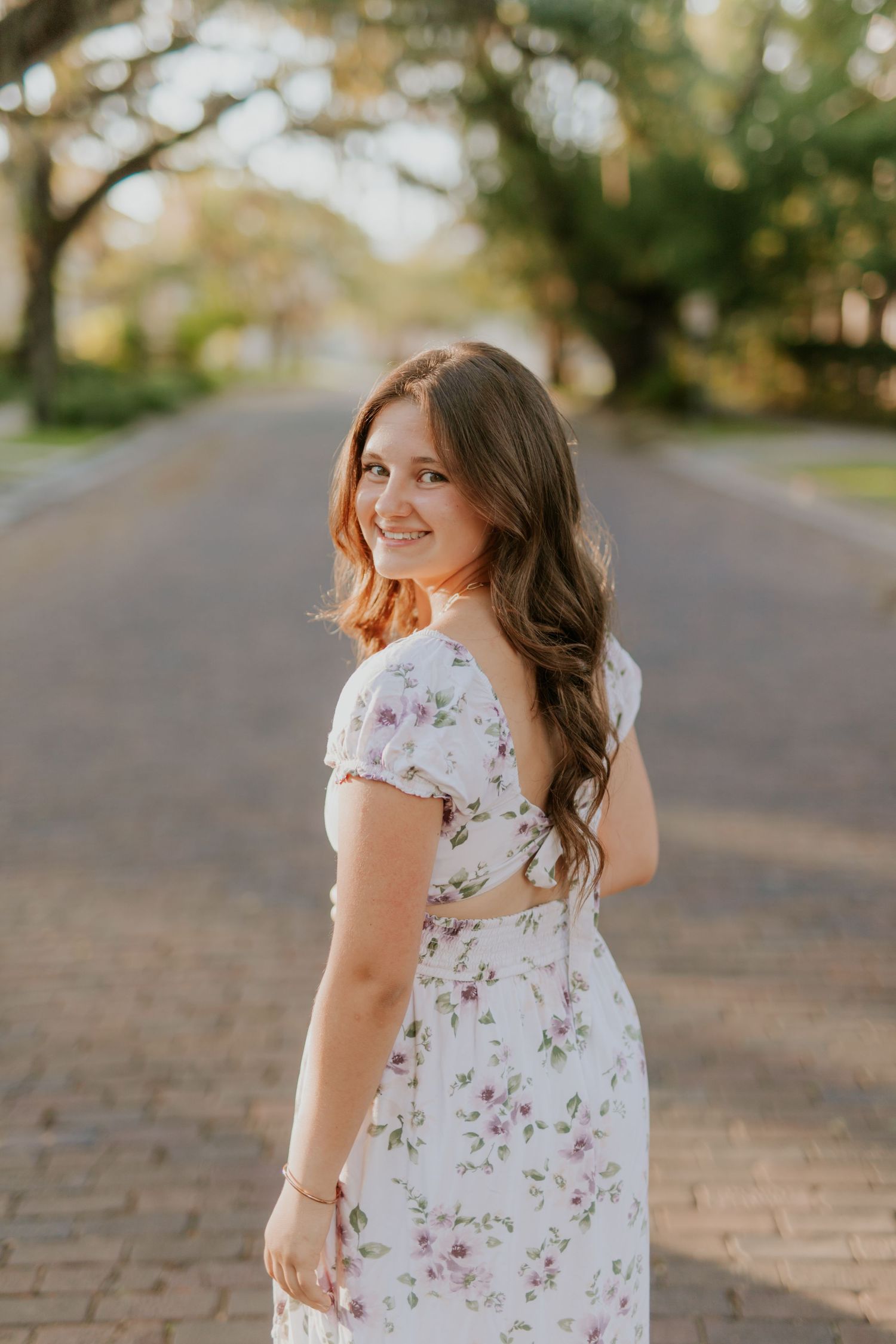 Young woman in floral white dress smiling over shoulder on tree-lined brick path in golden hour light.