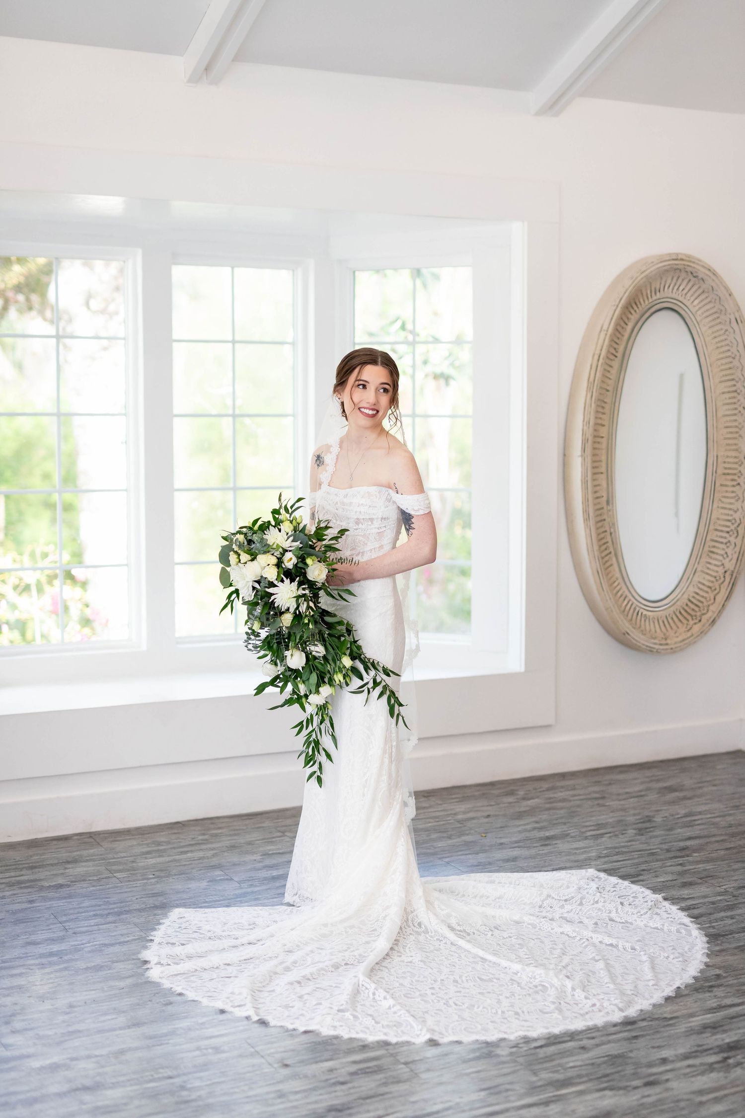 Bride in off-shoulder lace wedding gown with long train holds cascading white bouquet in bright white room.