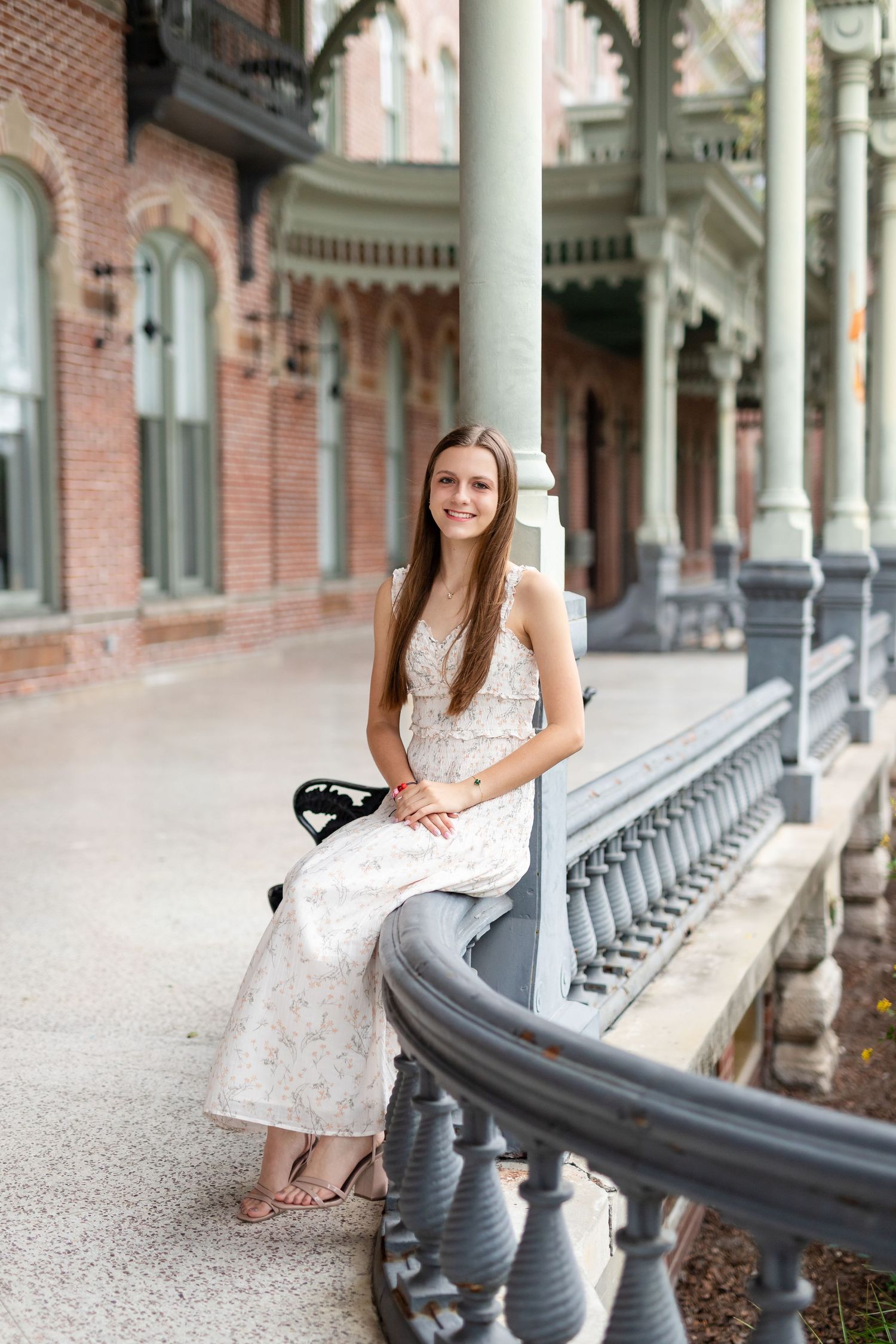 Elegant young woman in white floral dress sitting on railing of historic brick building porch at the University of Tampa.