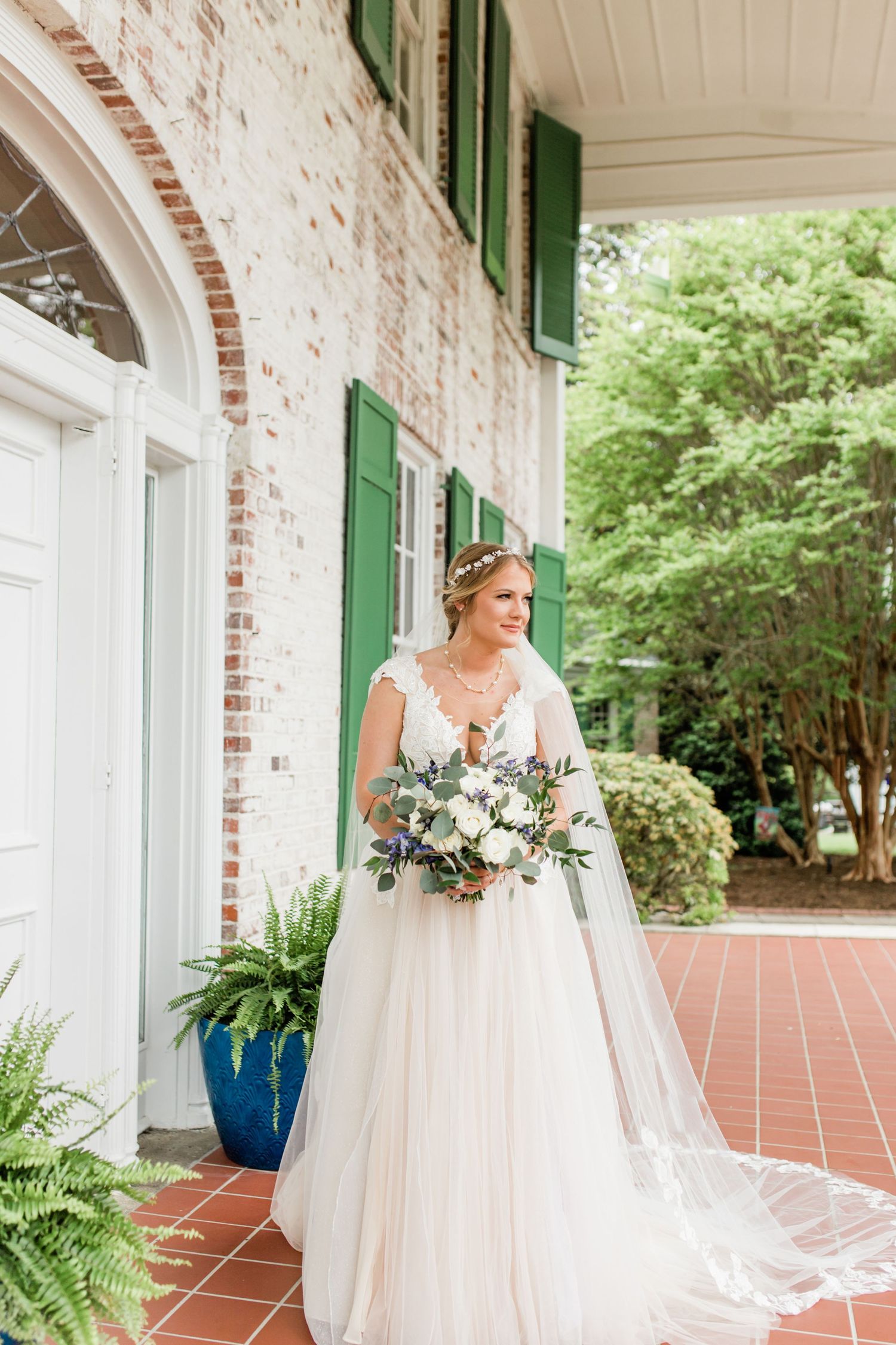 Bride in lace A-line gown with long veil holding bouquet on brick porch of historic venue.