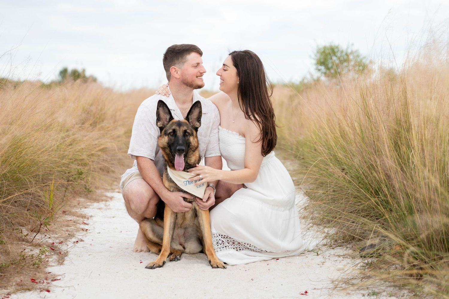 Couple sitting with their German Shepherd dog on a sandy path surrounded by tall golden grass at Honeymoon Island.