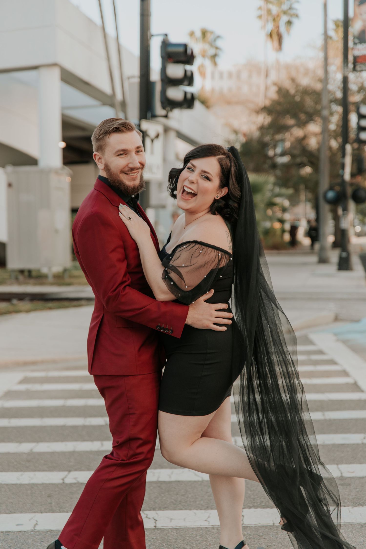 Laughing couple posing on city crosswalk, man in red suit, woman in black dress with veil.