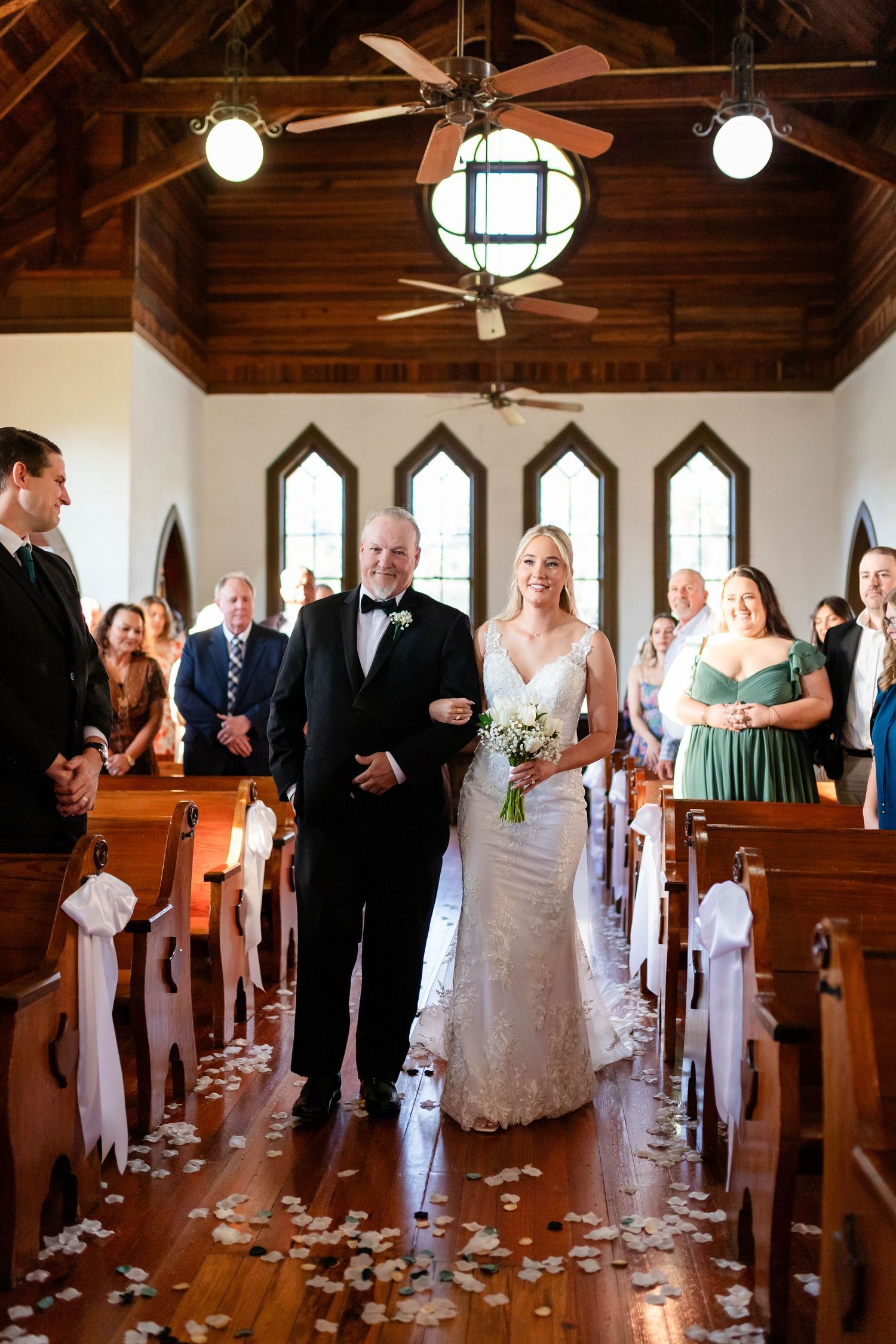 Bride in lace gown walking down the aisle with her father in a flower petal-lined church ceremony in Dunedin.