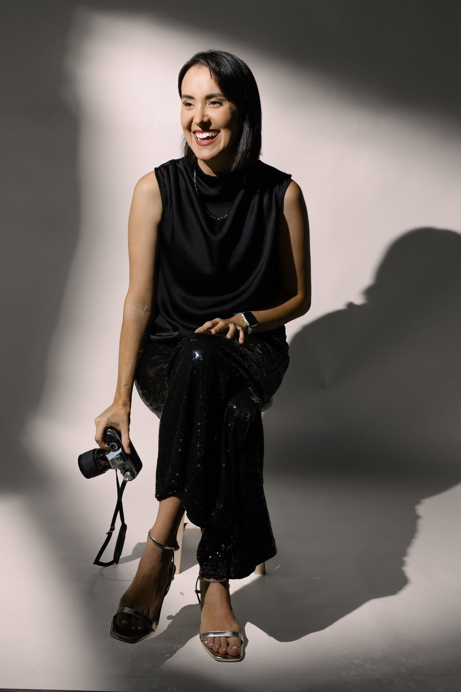 Smiling woman in black sleeveless dress holding a camera, seated against a light gray studio background.