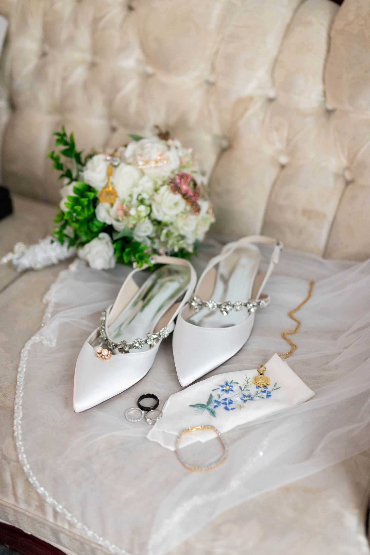 White bridal heels with crystal embellishments displayed on a tufted sofa beside a floral bouquet.