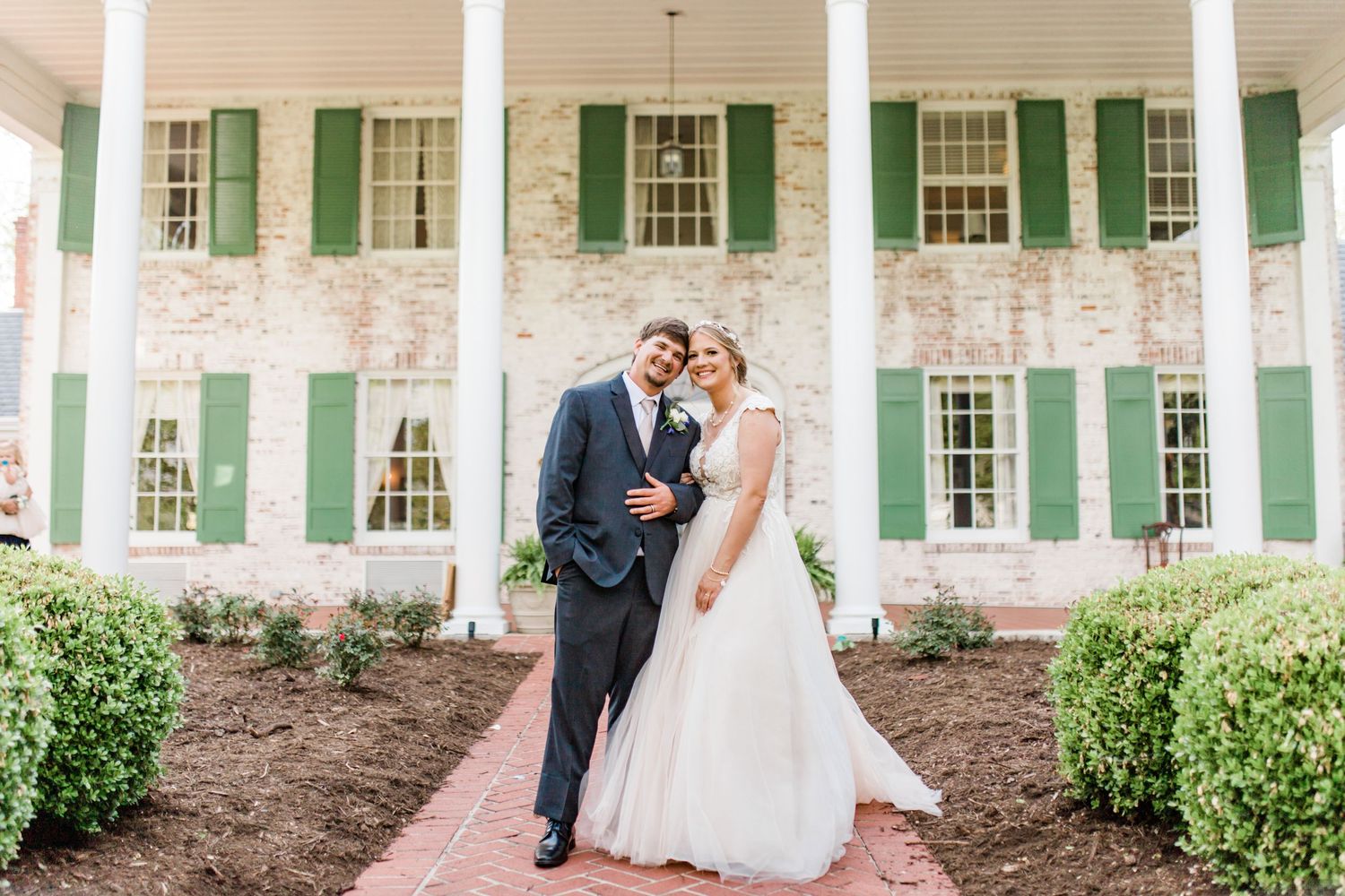 Bride and groom pose together in front of a historic brick building with green shutters on their wedding day.