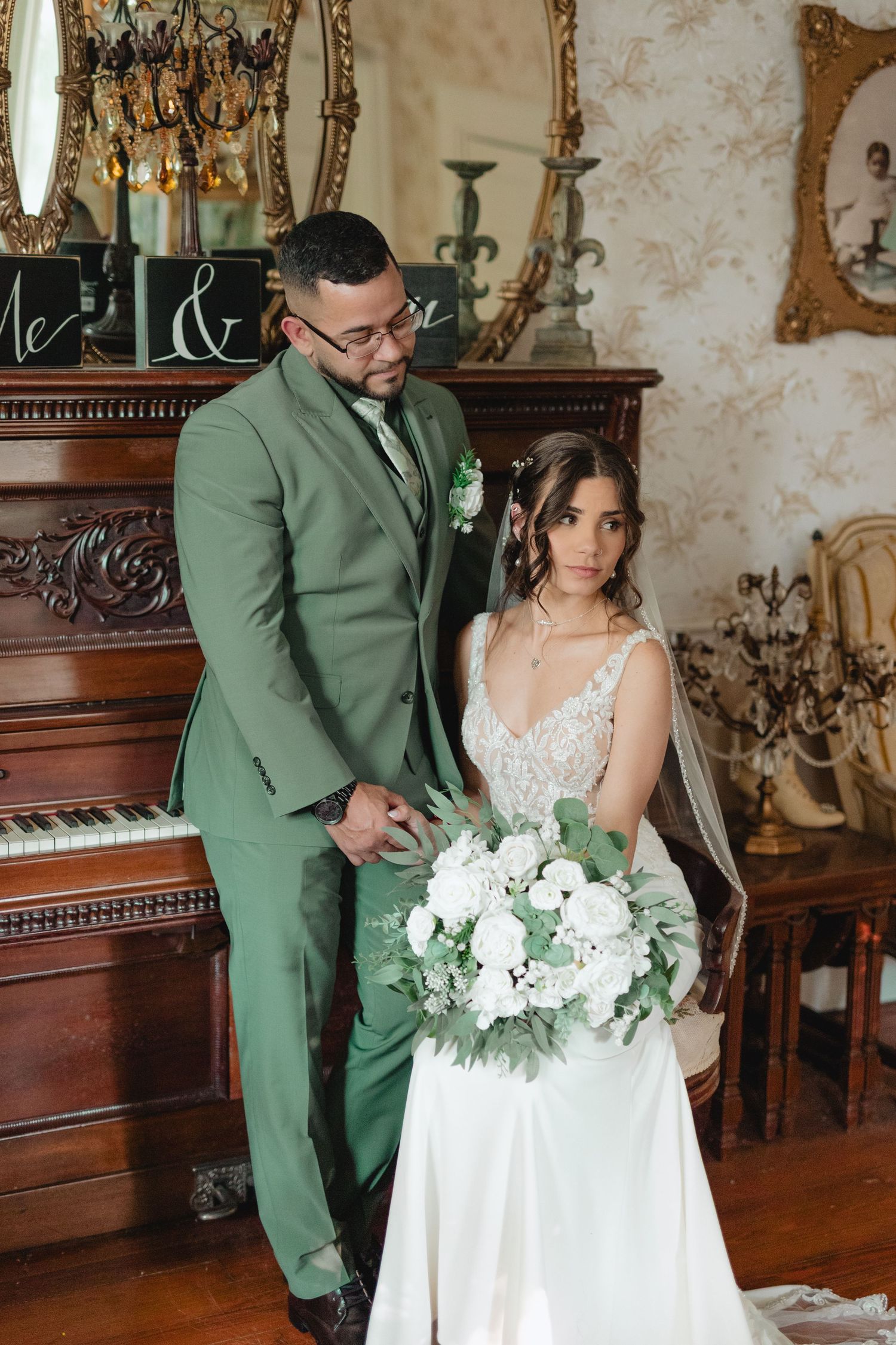 Groom in green suit stands beside seated bride holding white bouquet in front of ornate vintage piano indoors.