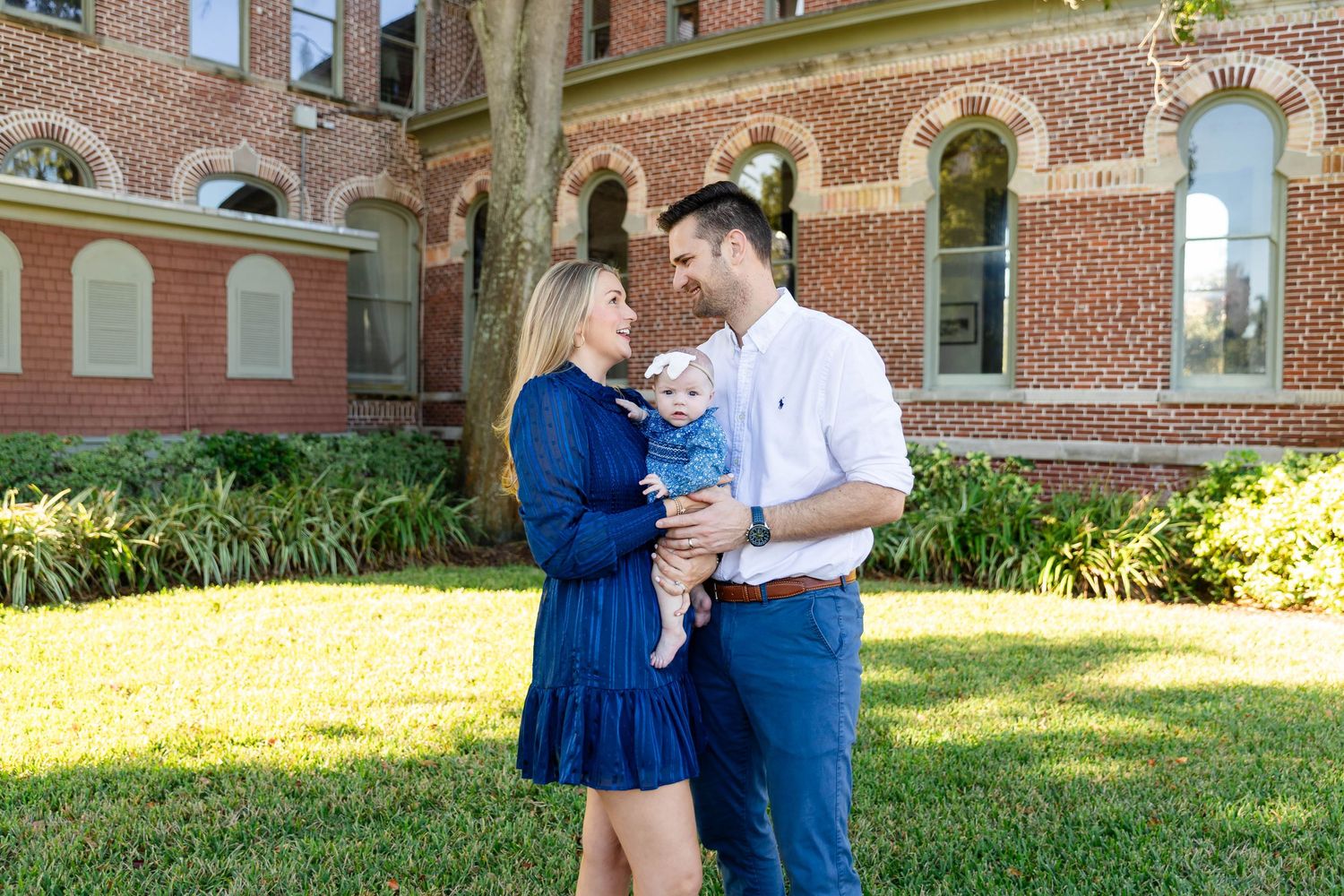 Young couple in blue outfits holds baby boy while standing on green lawn near brick building at the University of Tampa.