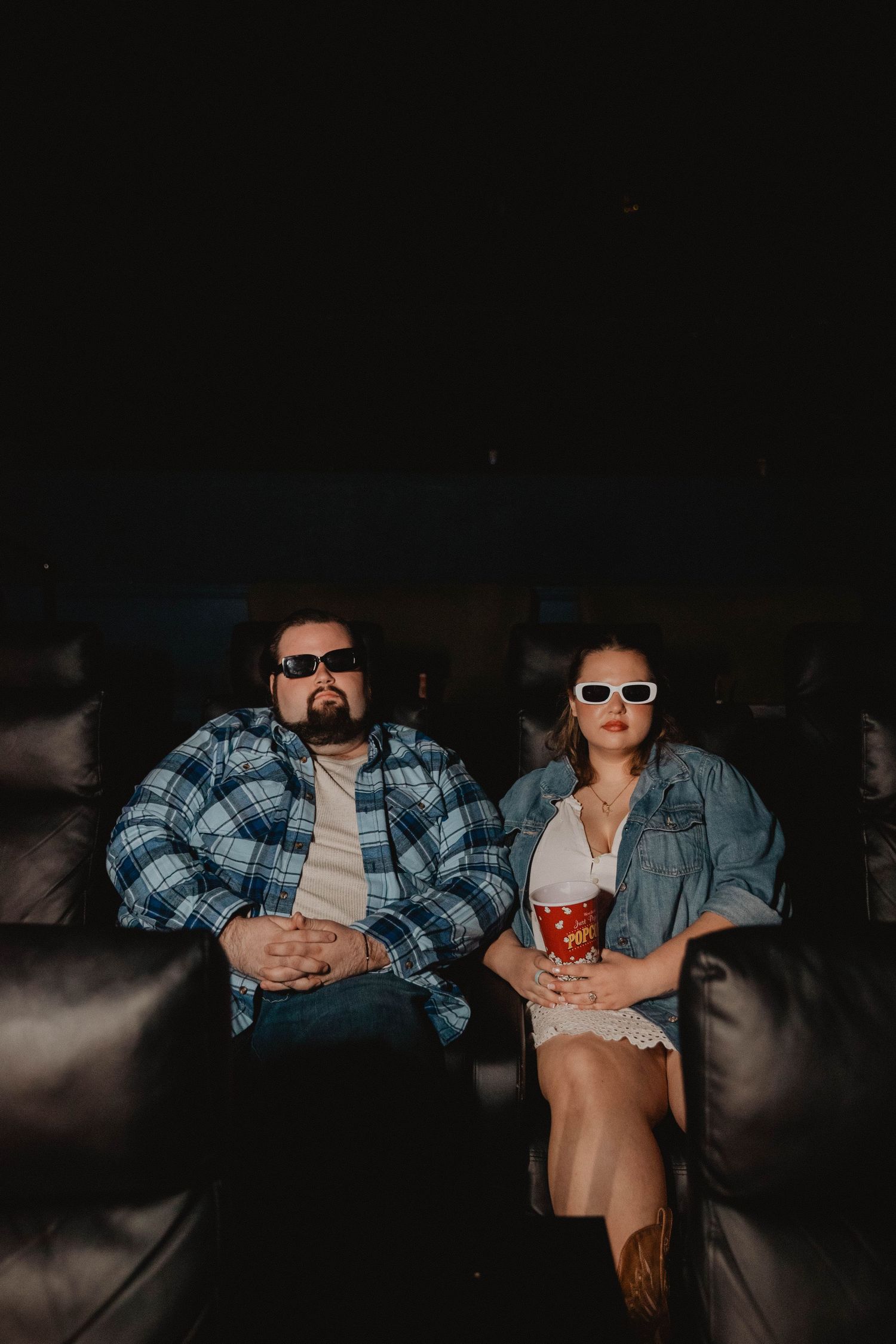 Couple wearing sunglasses sitting on a leather couch watching a movie with popcorn in a dark movie theatre.