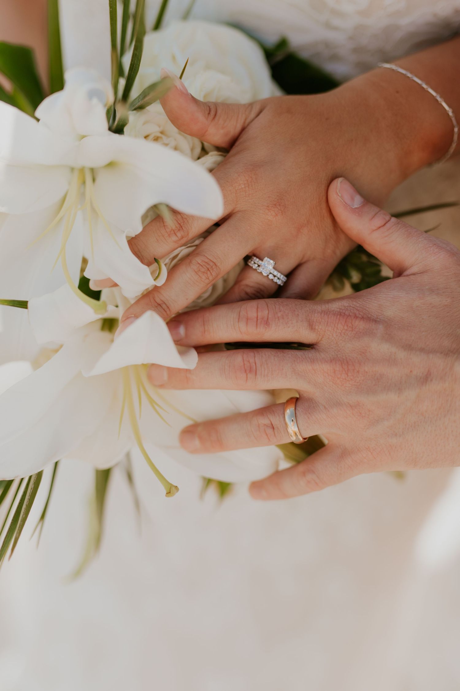 Close-up of bride and groom hands showing wedding rings resting on white bouquet.