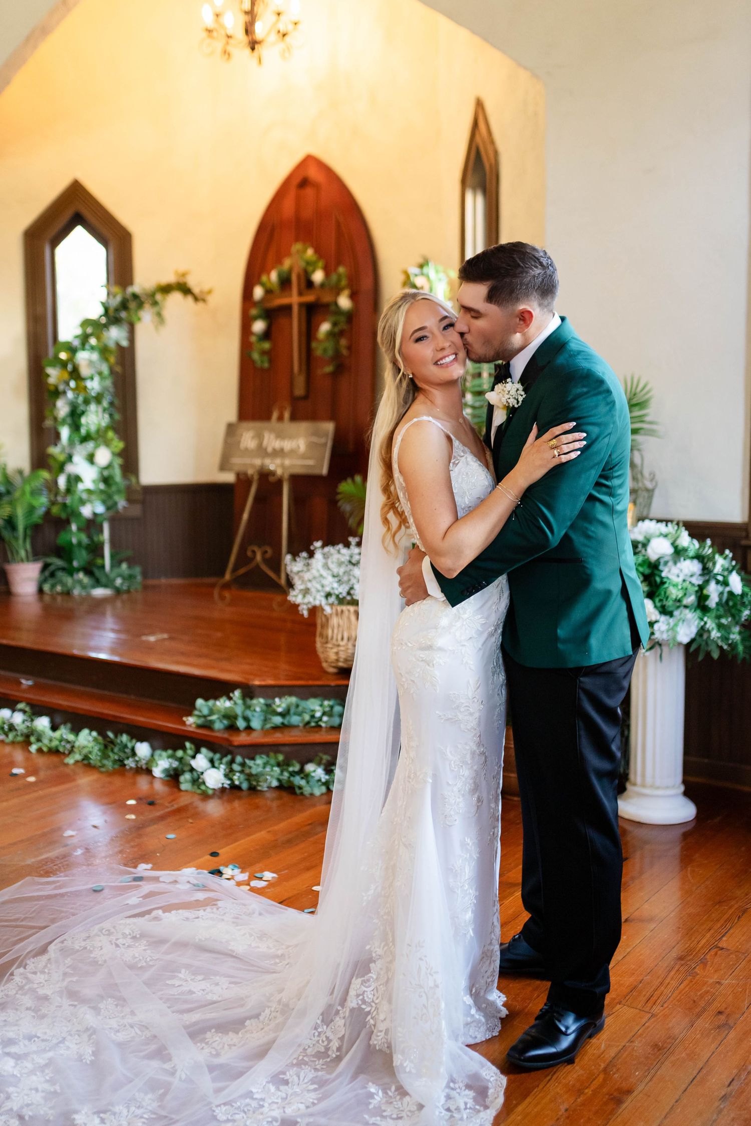 Newlyweds embracing and smiling in a charming chapel adorned with white floral arrangements and a green velvet tuxedo in Dunedin.