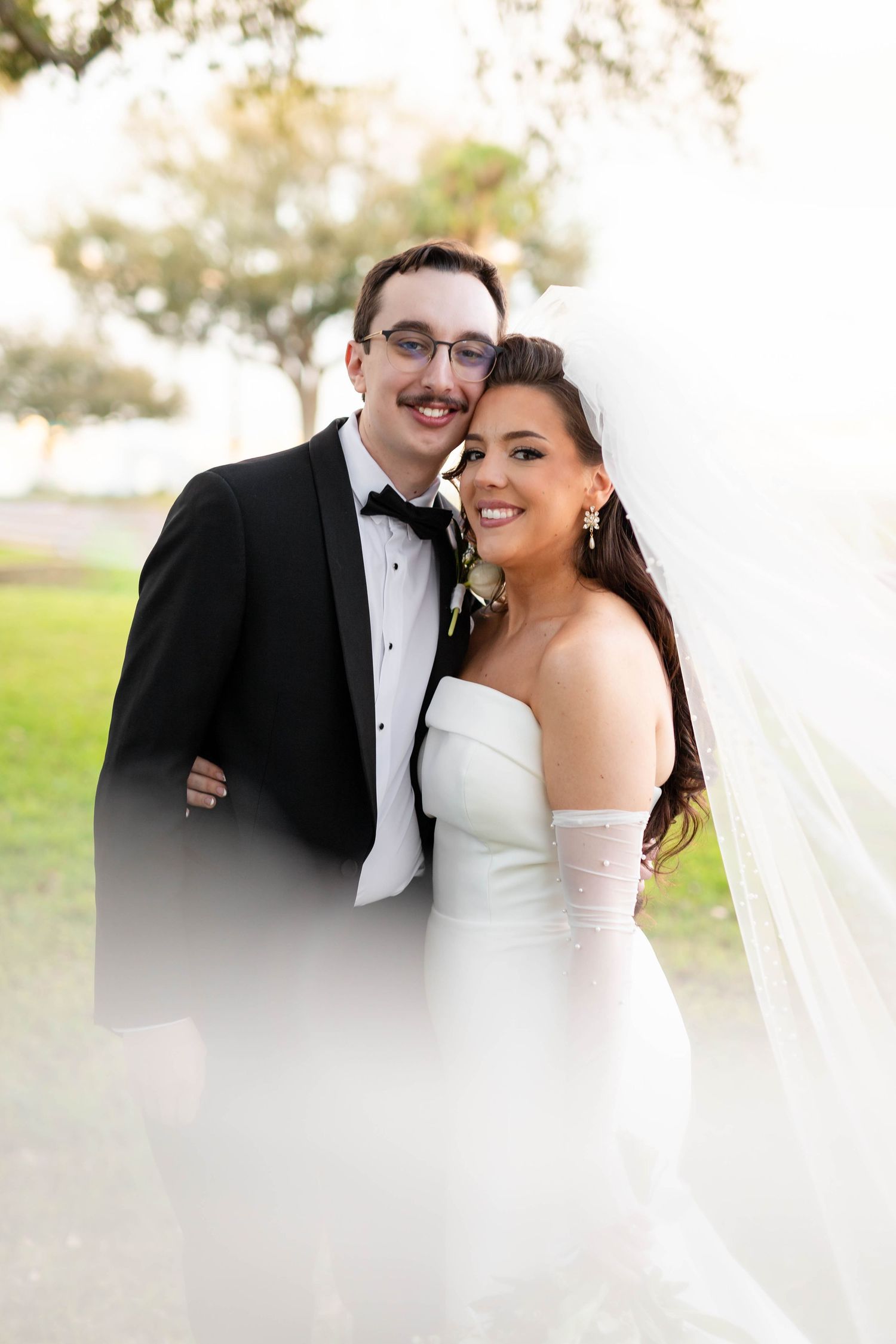 Smiling bride and groom pose together outdoors, bride in strapless white gown with veil, groom in black tuxedo on Boyshore Blvd in Tampa.