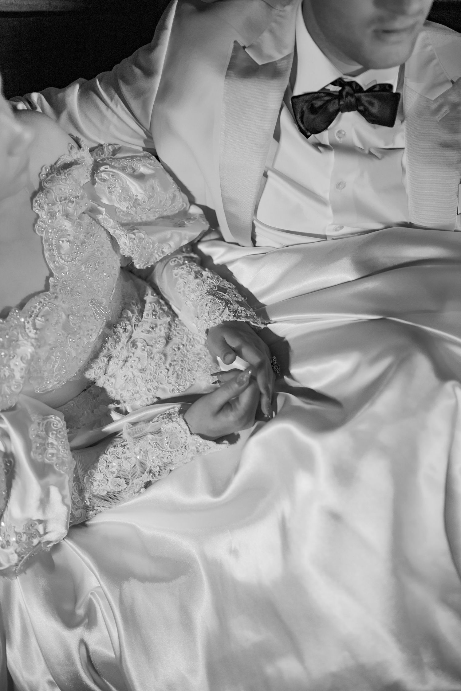 Black and white photo of bride and groom holding hands, showcasing lace wedding dress and groom's bow tie.