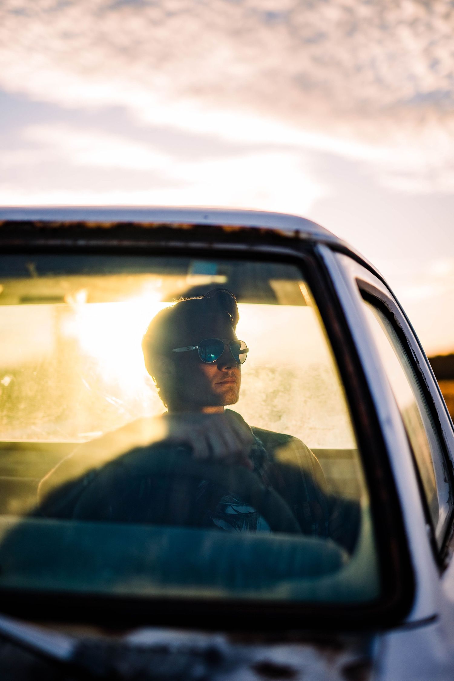 Young man in sunglasses seen through car window with golden sunset light reflecting off glass.