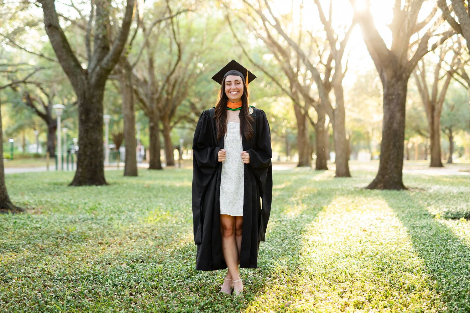 Female graduate in black cap and gown walking through a sunlit tree-lined park pathway smiling at the University of South Florida.