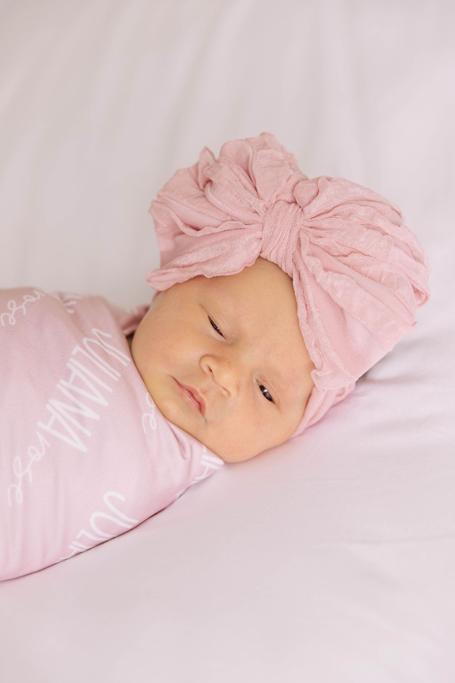 Newborn baby girl swaddled in pink personalized blanket wearing a soft pink bow turban hat.