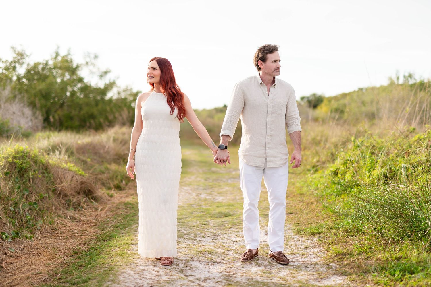 Couple holding hands and strolling down a rustic sandy path with lush greenery in the background at Honeymoon Island.