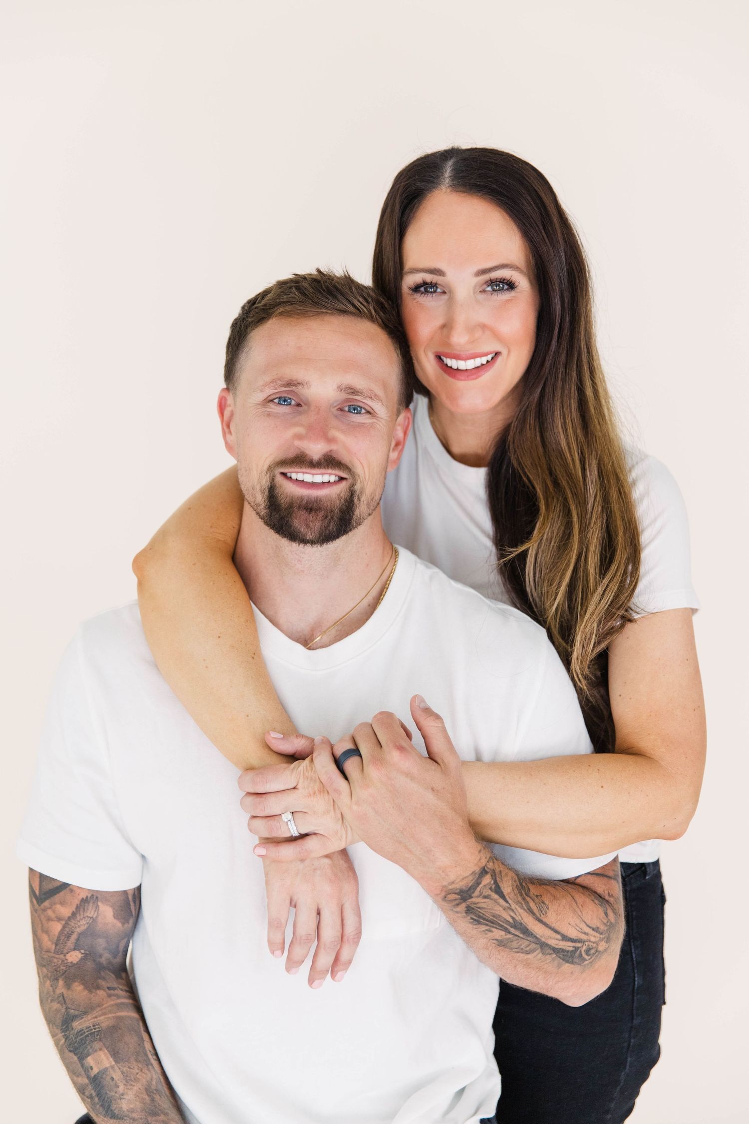 Smiling couple in white tees, tattooed man with woman hugging him from behind.