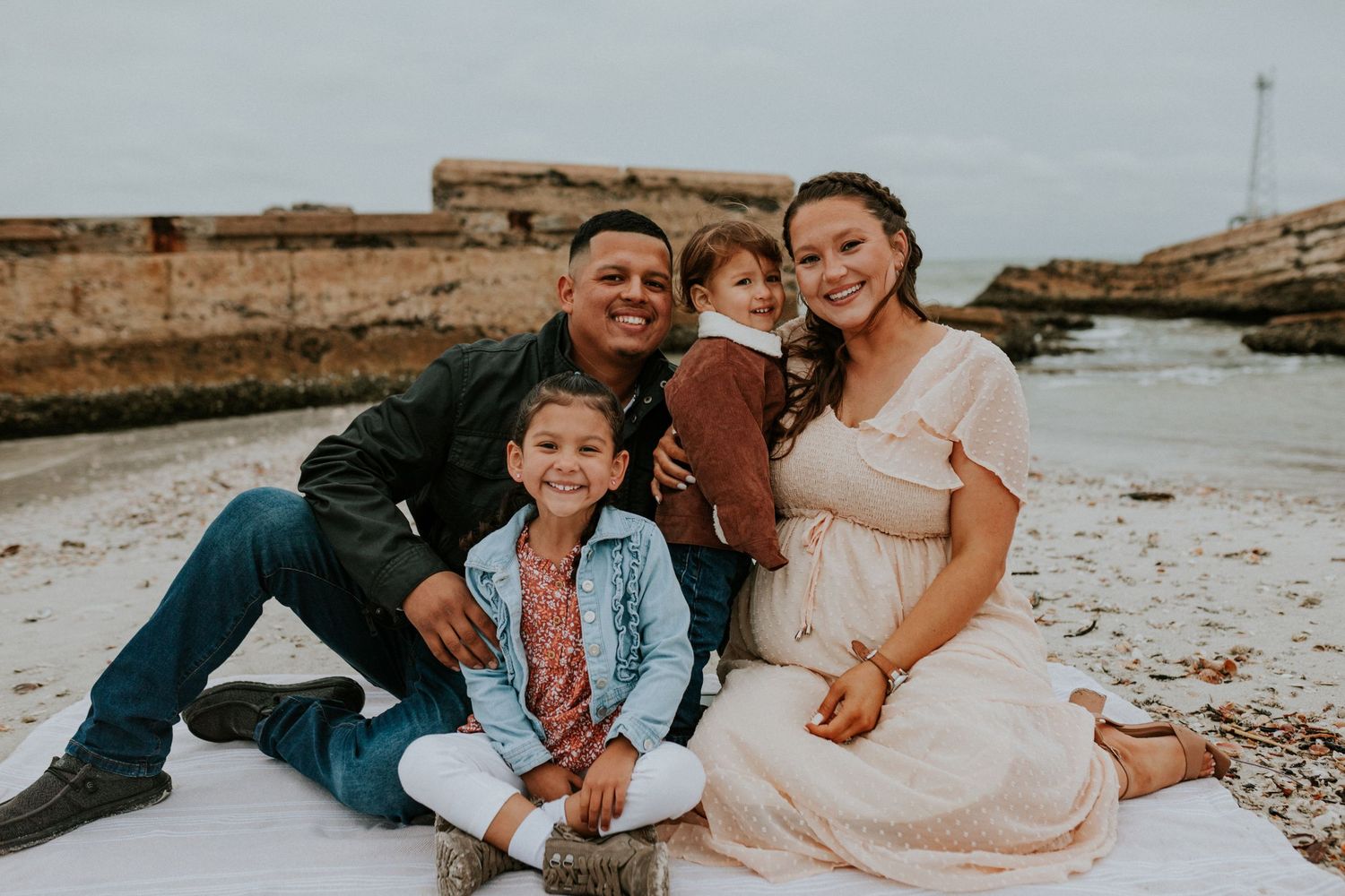 A happy family of four sits together on a rocky beach, smiling for a casual outdoor portrait.