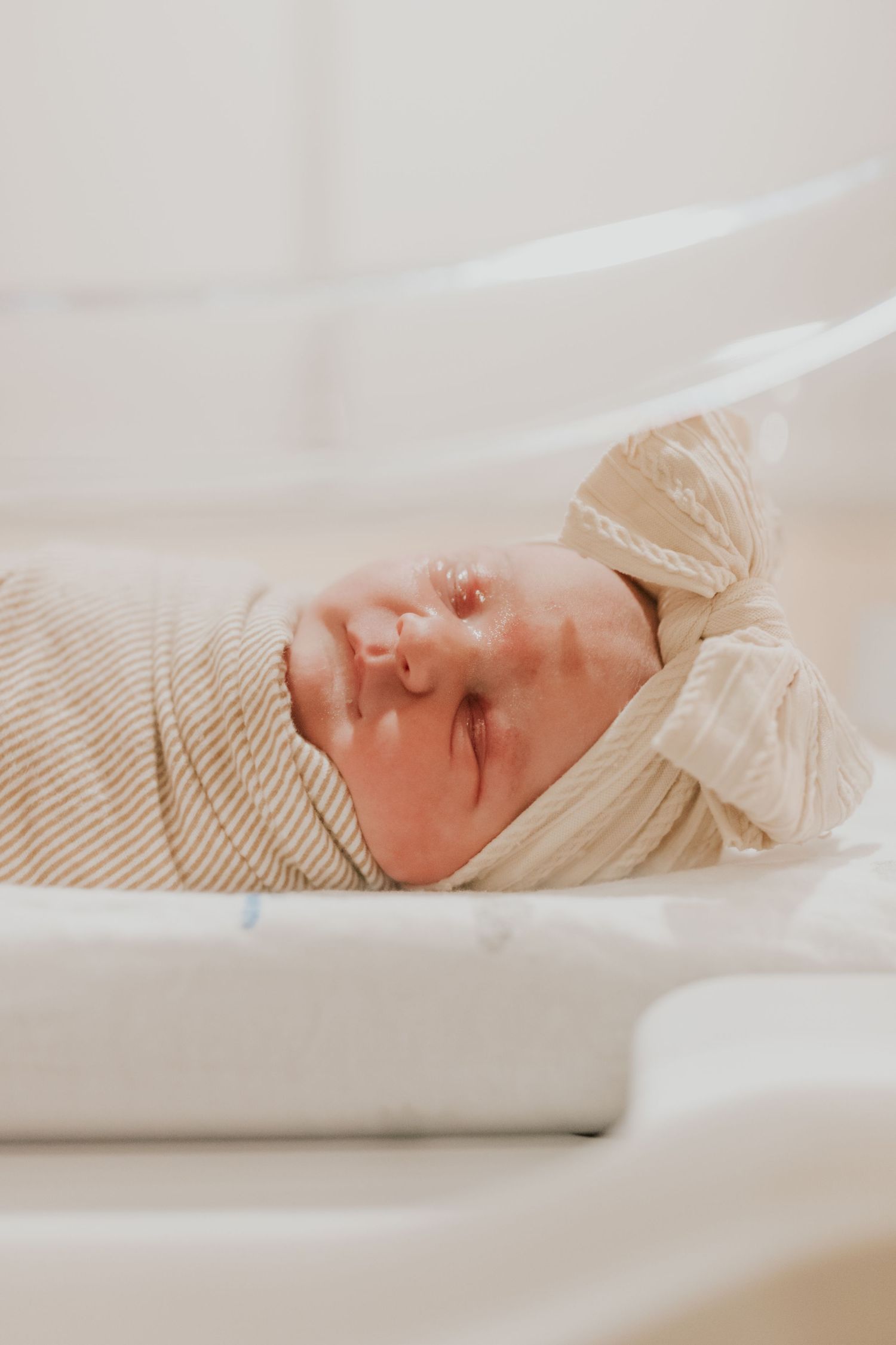 Peaceful newborn baby swaddled in striped blanket with bow headband sleeping in hospital bassinet.
