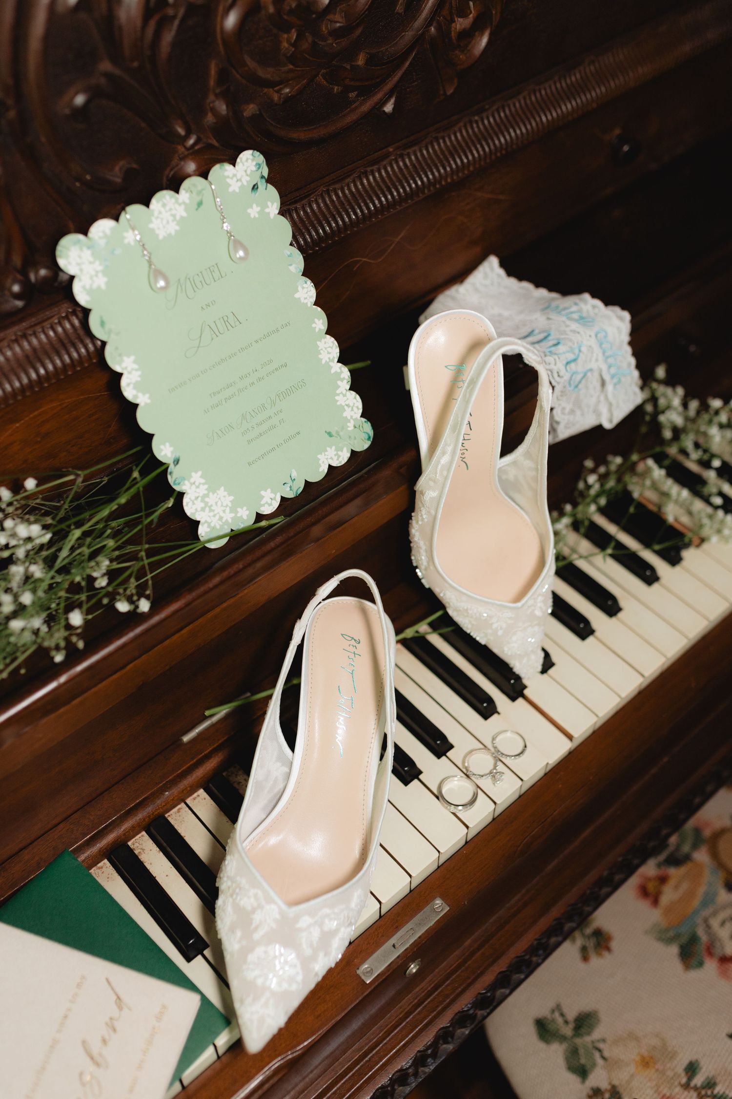 White lace bridal heels and mint green wedding invitation displayed on antique piano keys with baby's breath flowers.