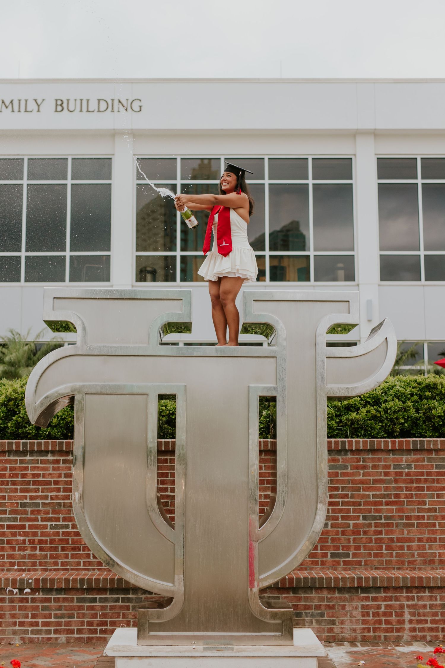 Graduate in white dress stands on Tampa University TU statue, spraying champagne to celebrate commencement.
