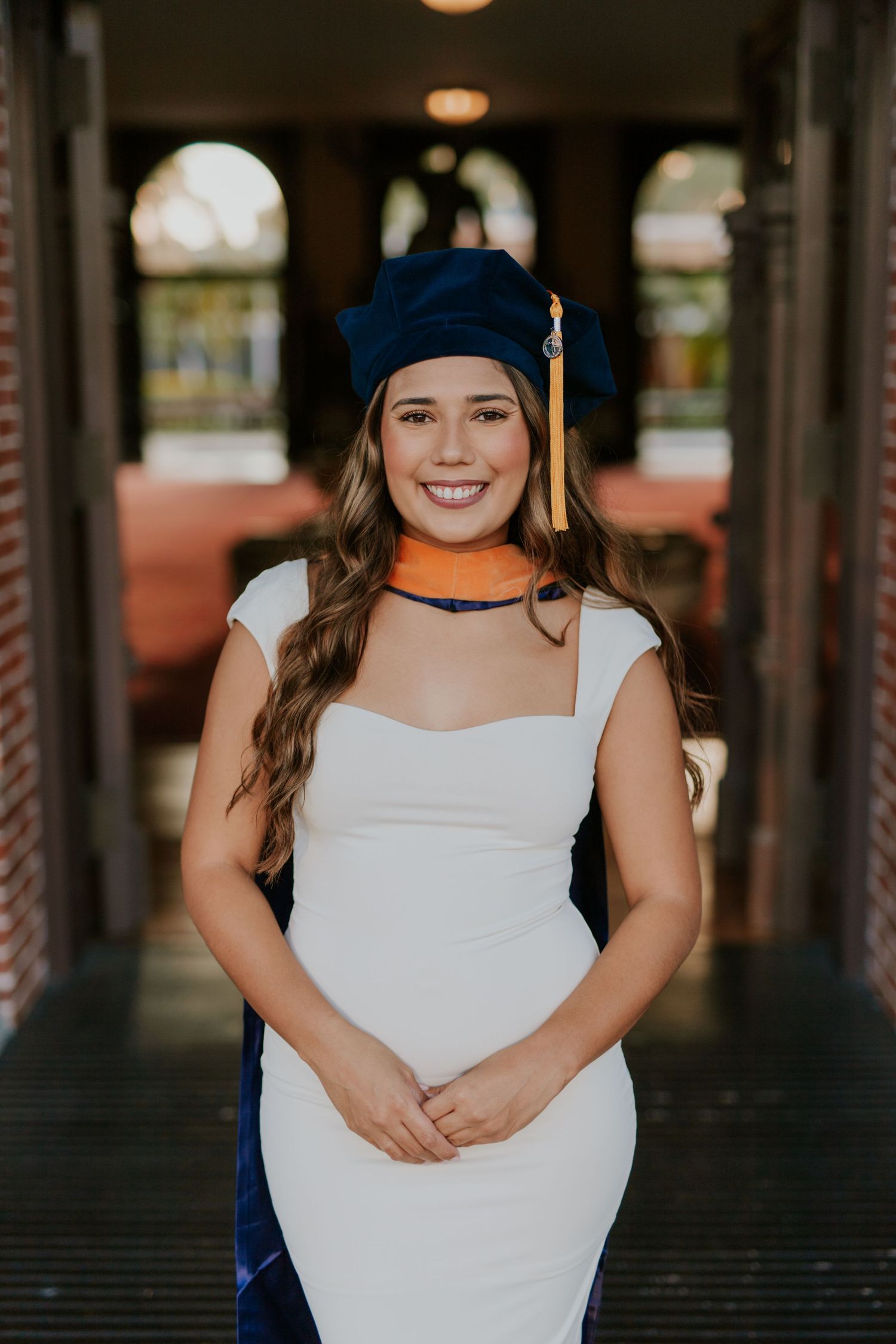 Smiling graduate in white dress and navy blue cap with gold tassel poses outdoors for graduation portrait.