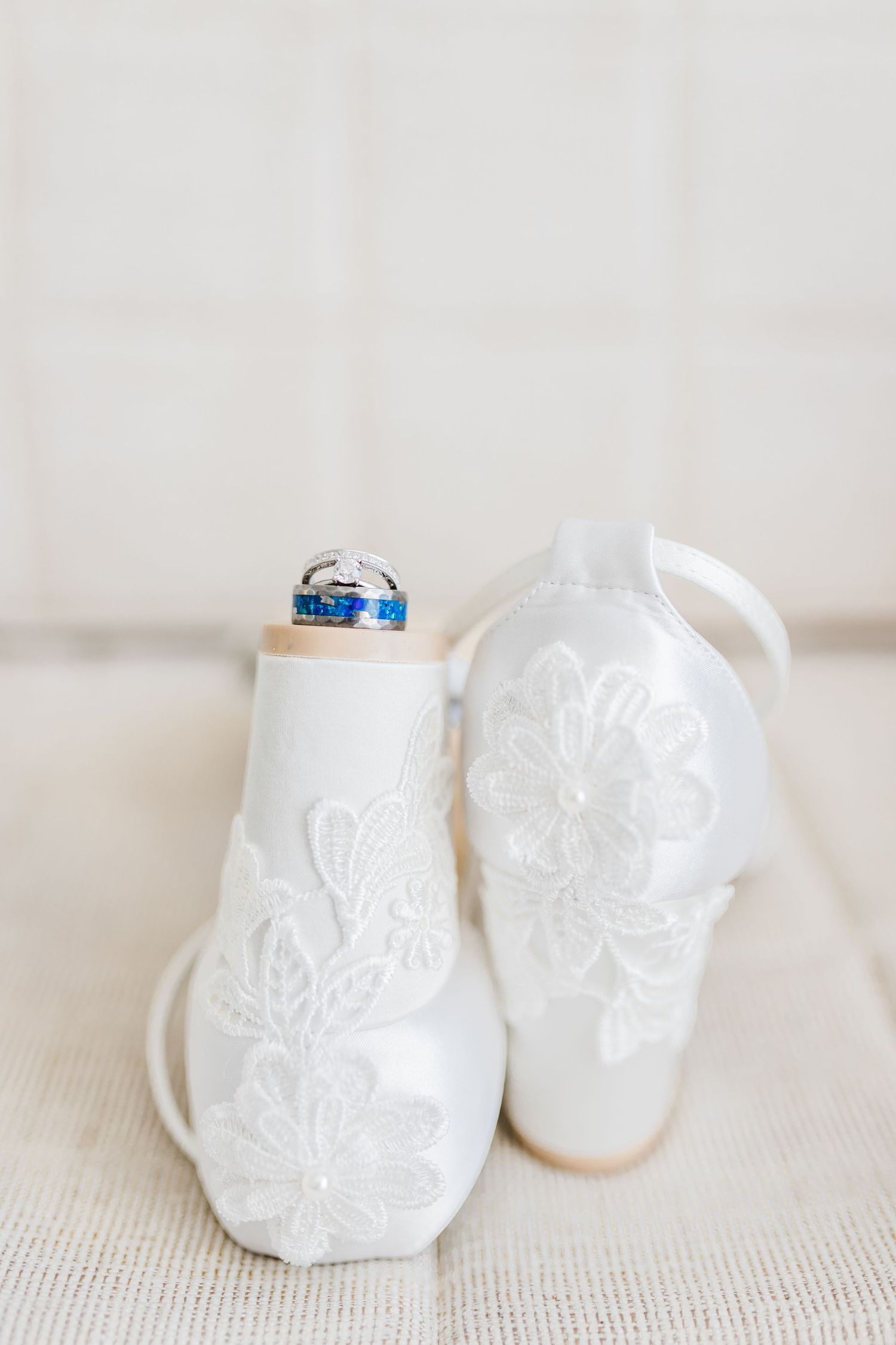 White lace bridal heels with blue and silver wedding rings resting between the shoes.