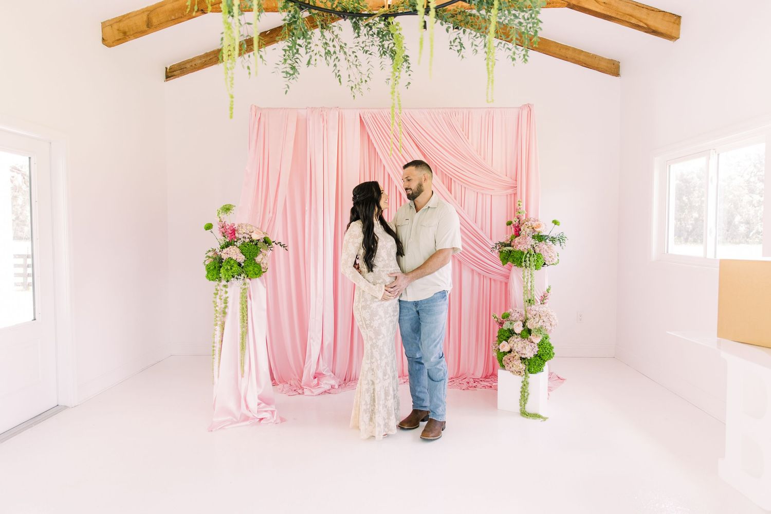 Expecting couple shares intimate moment before pink draped backdrop with lush floral arrangements in bright venue at the Colonial at Sunset.