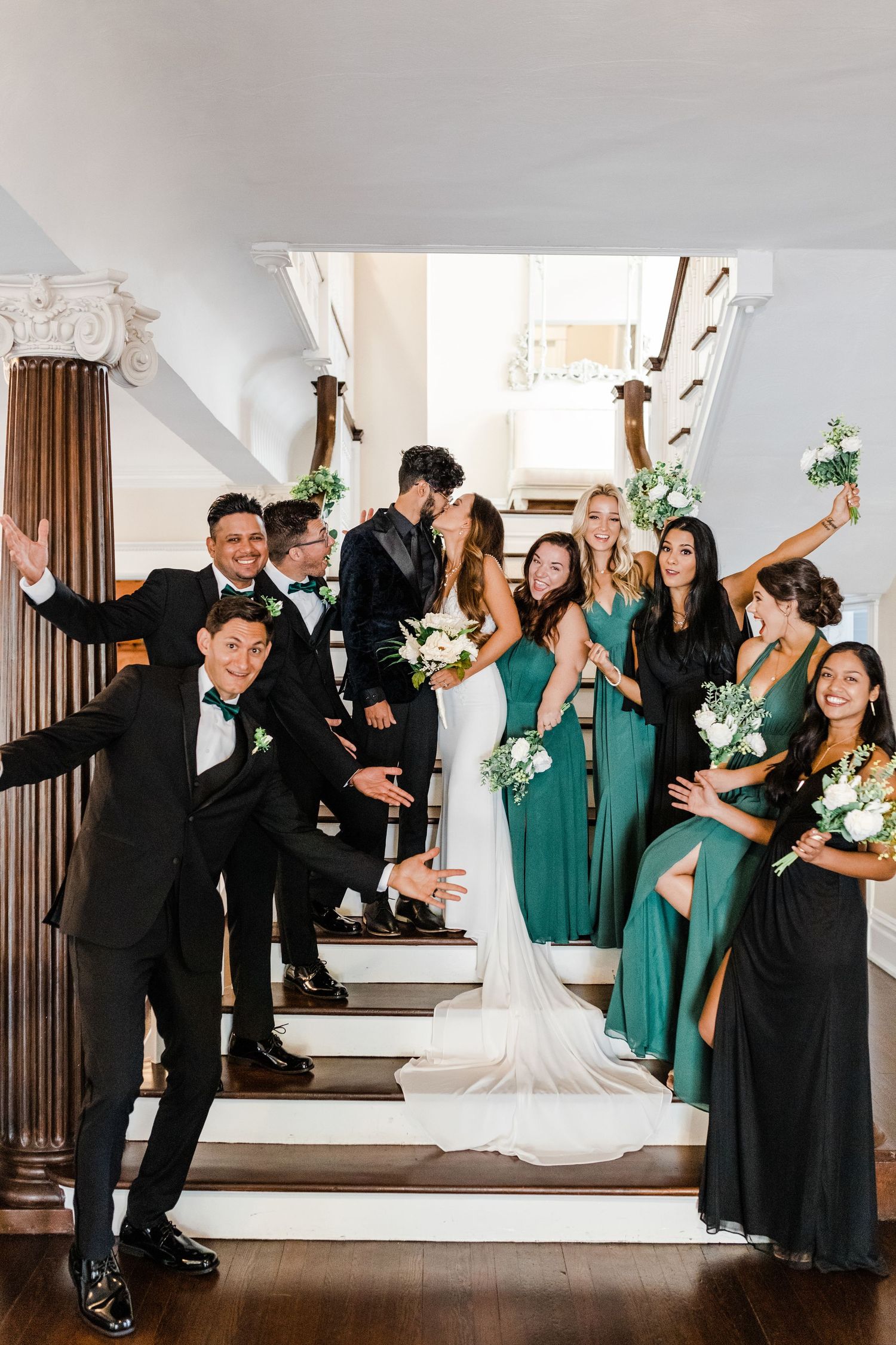 Wedding party poses on elegant staircase, bridesmaids in emerald green gowns, groomsmen in black tuxedos, bride and groom kissing, The Orlo.