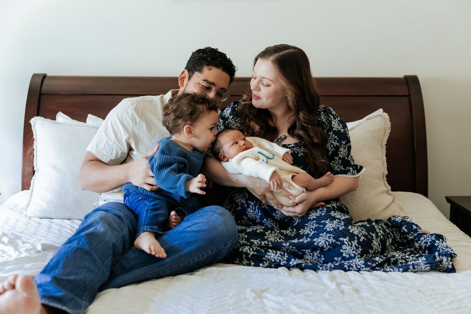 Family of four with toddler and newborn cuddles together on a white bed at home.