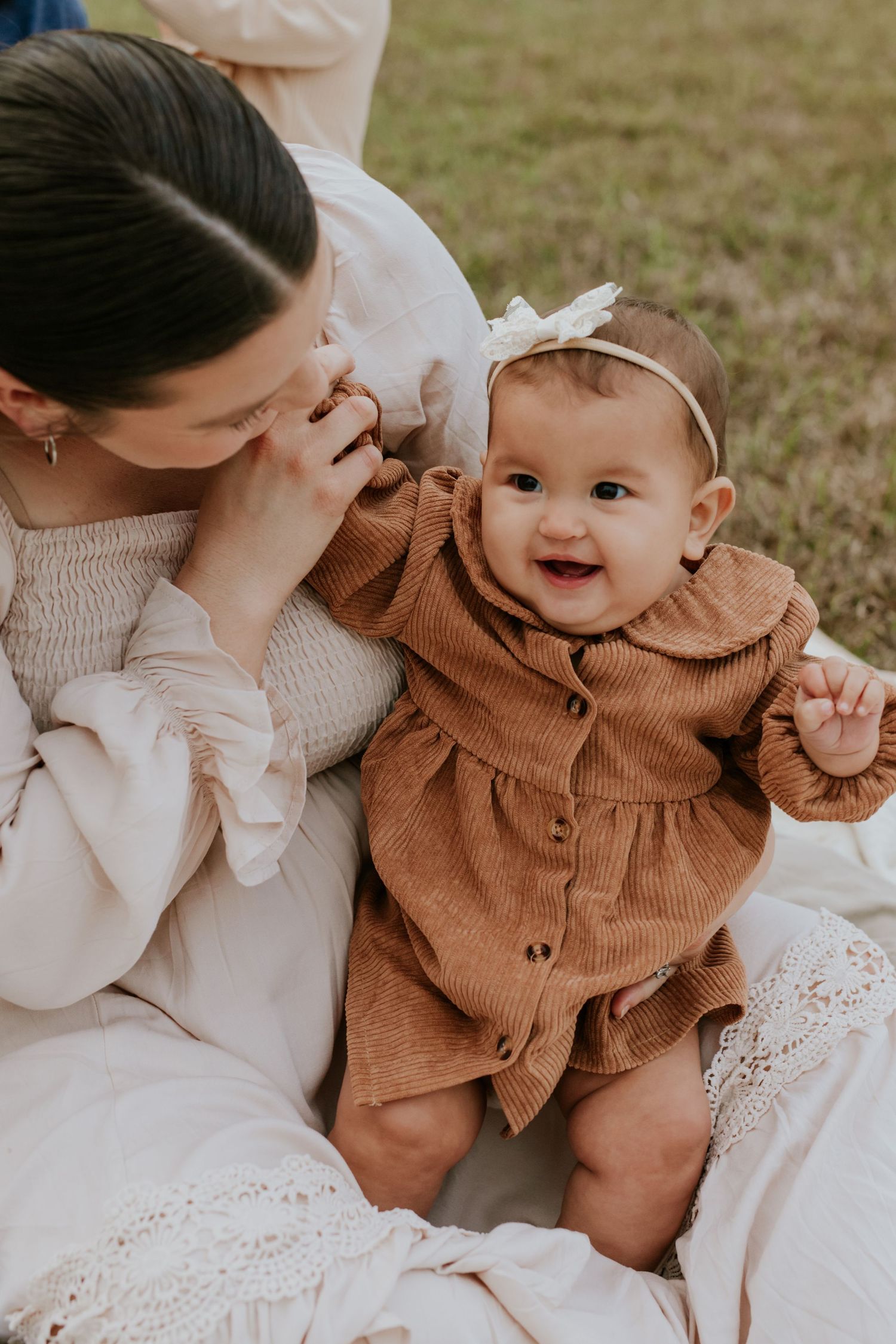 Smiling baby in brown corduroy dress with bow headband sitting with mother outdoors.