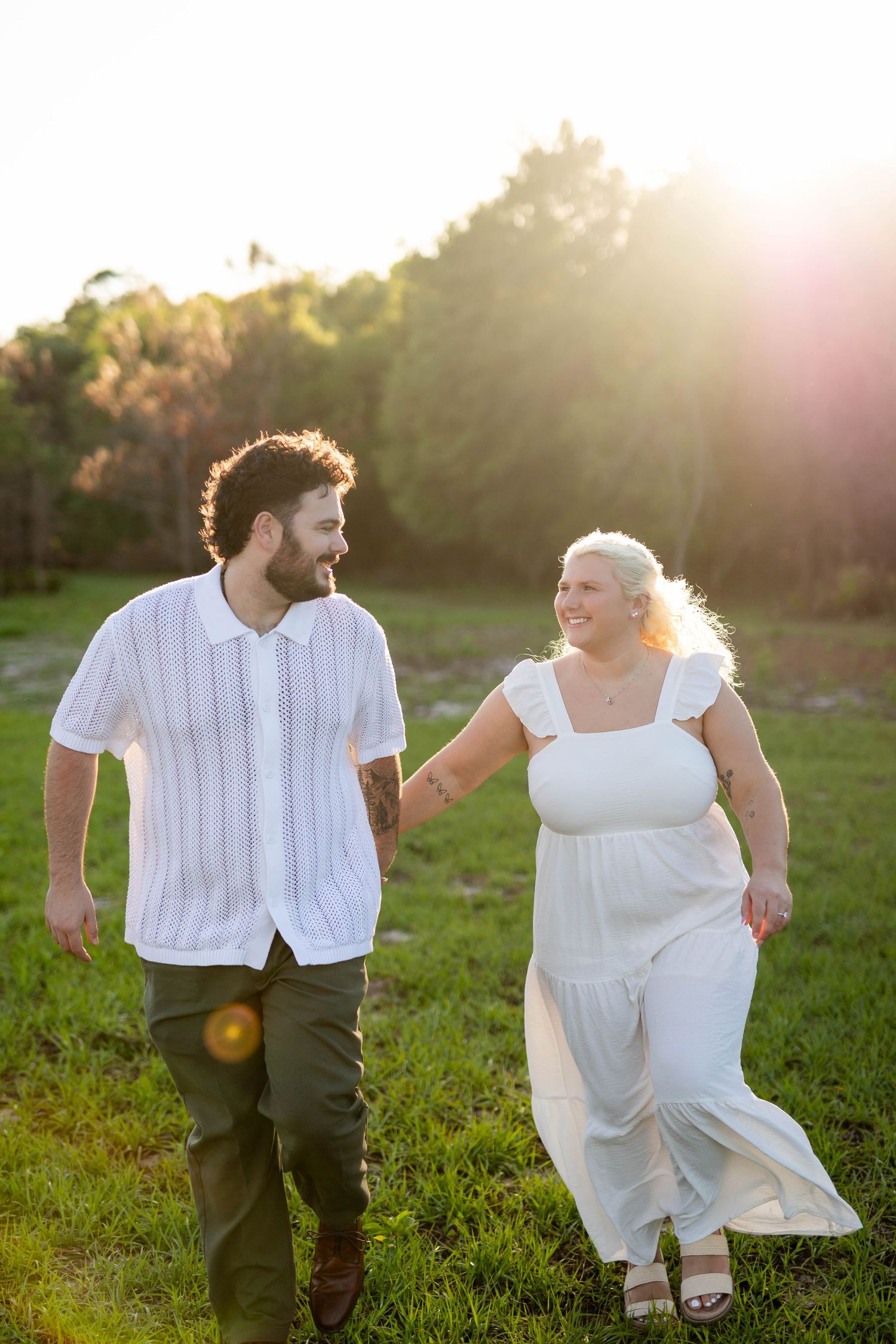Happy couple holding hands and walking through a sunny green field, dressed in white and olive tones.