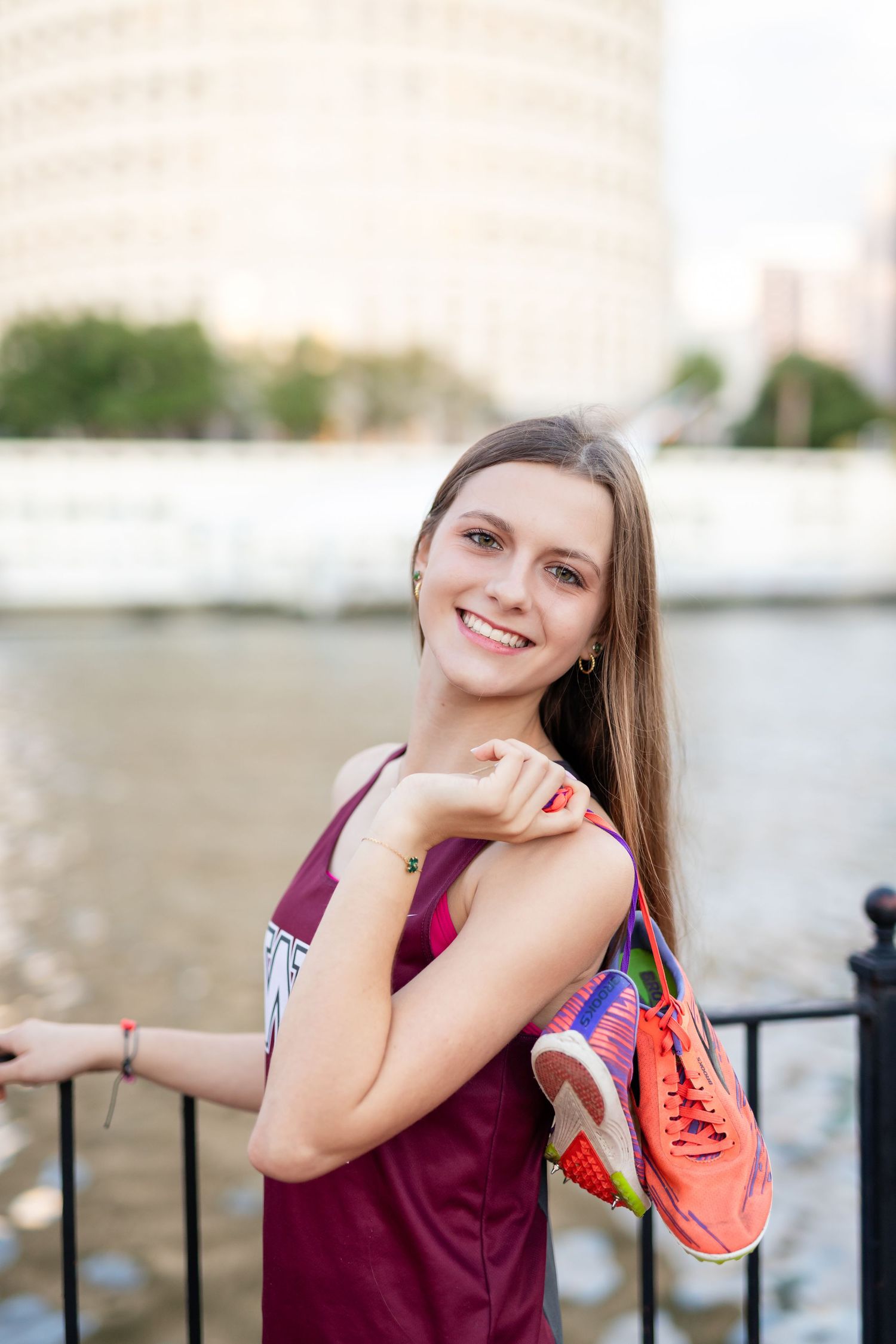 Smiling young woman in burgundy athletic top holding colorful sneakers by a city waterfront on a sunny day at the University of Tampa.