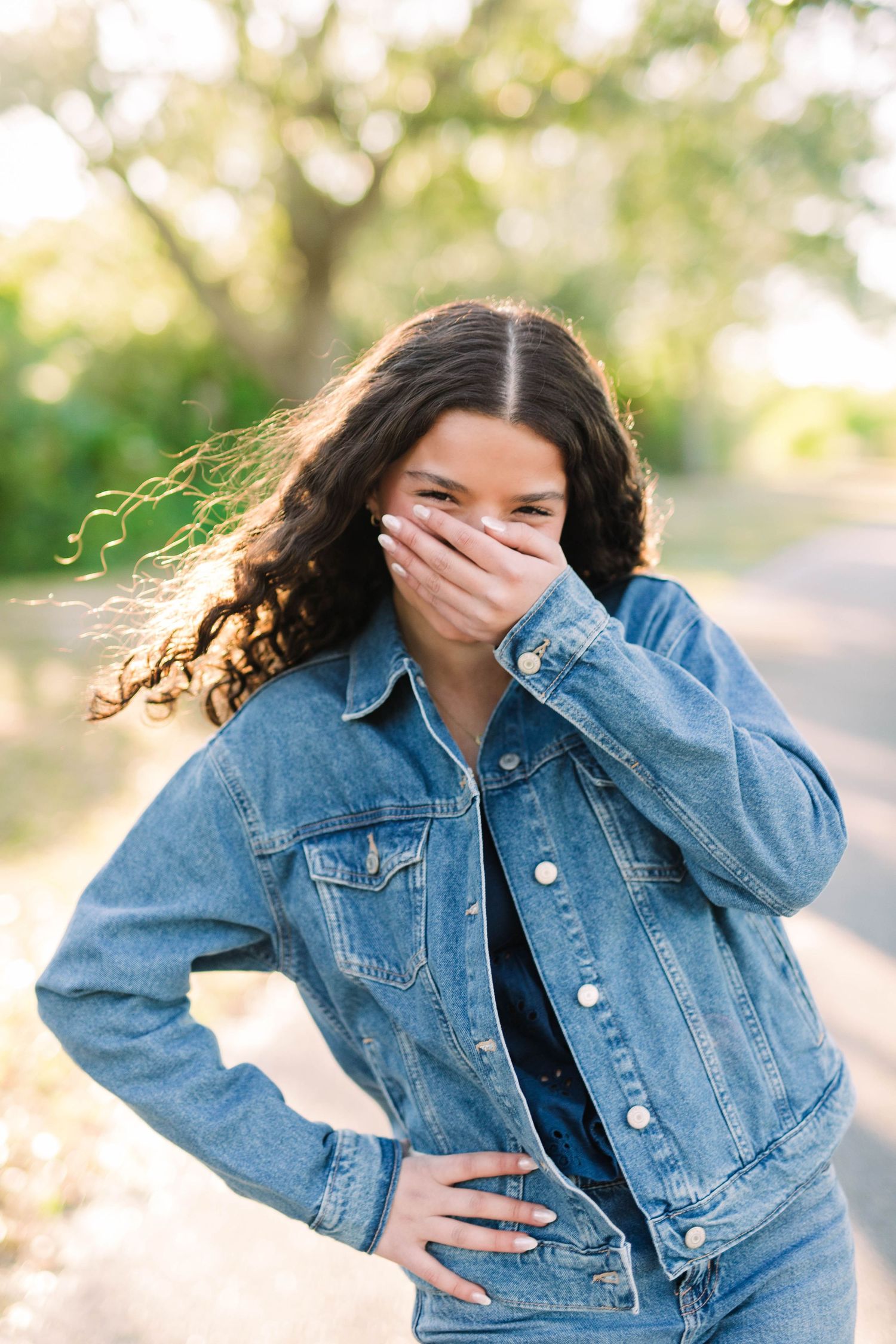 Young woman in denim jacket laughs while covering her mouth outdoors on a sunny day at Cypress Pointe Park.