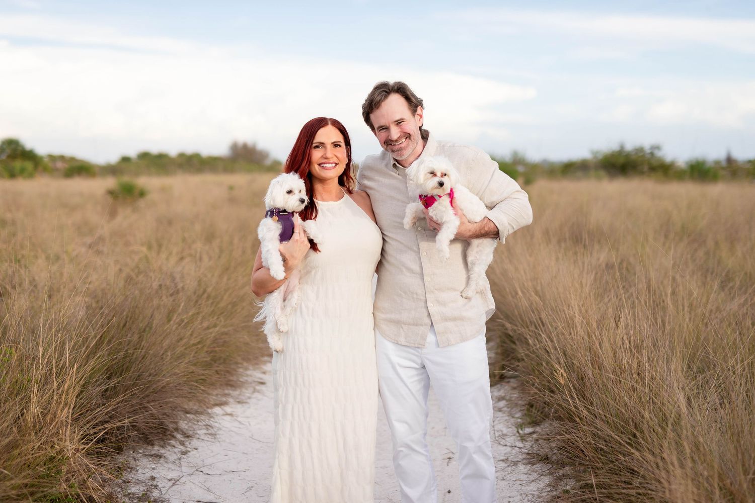 Smiling couple in white outfits holding small white dogs in a scenic marsh field at Honeymoon Island.