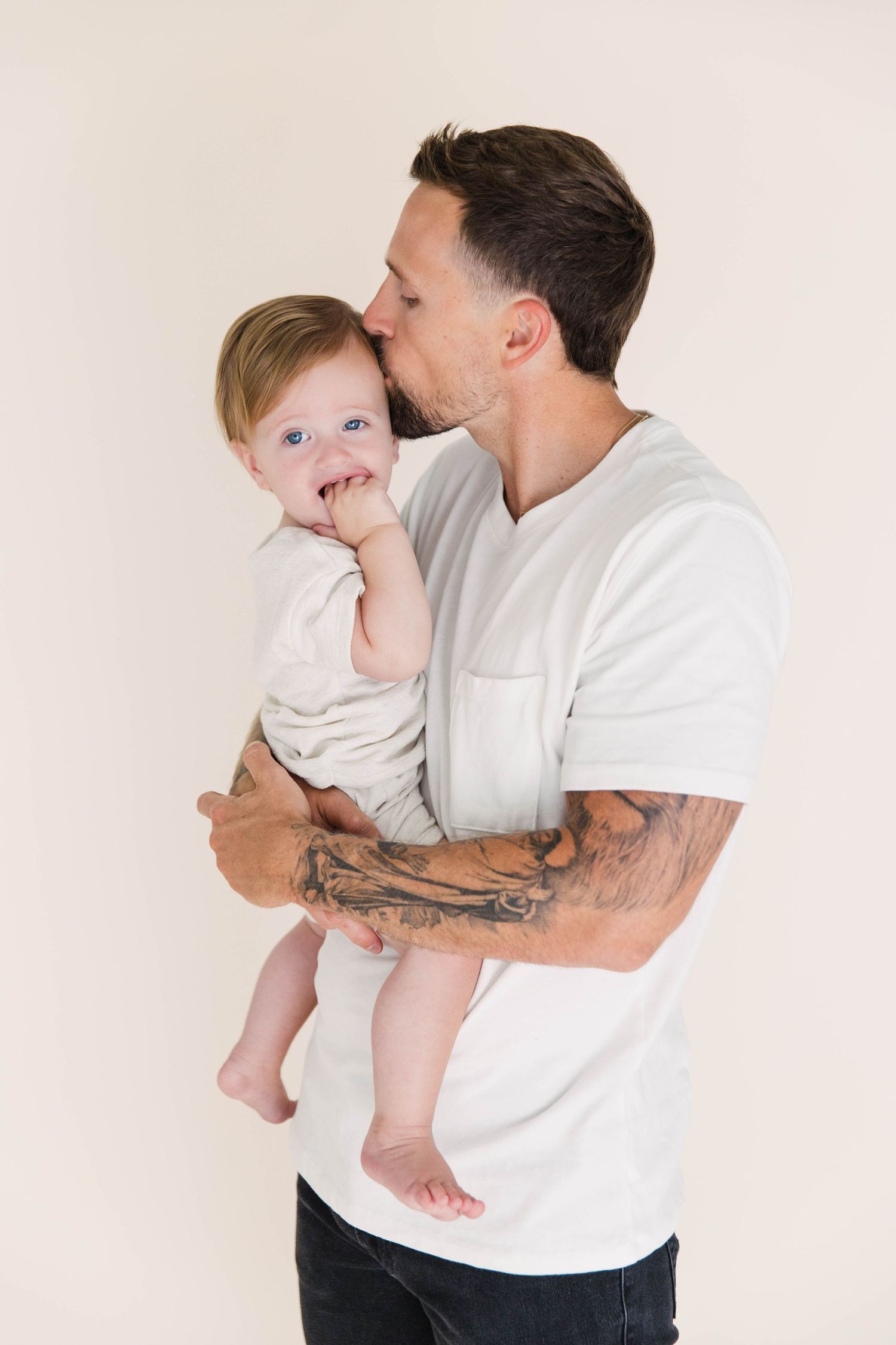 Tattooed father kissing adorable blue-eyed baby on cream studio background.