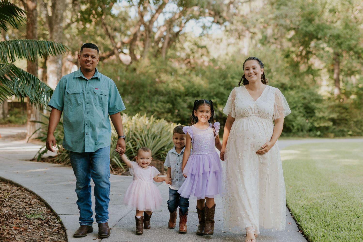 Pregnant woman in white dress walks with husband and two young children on a garden path.