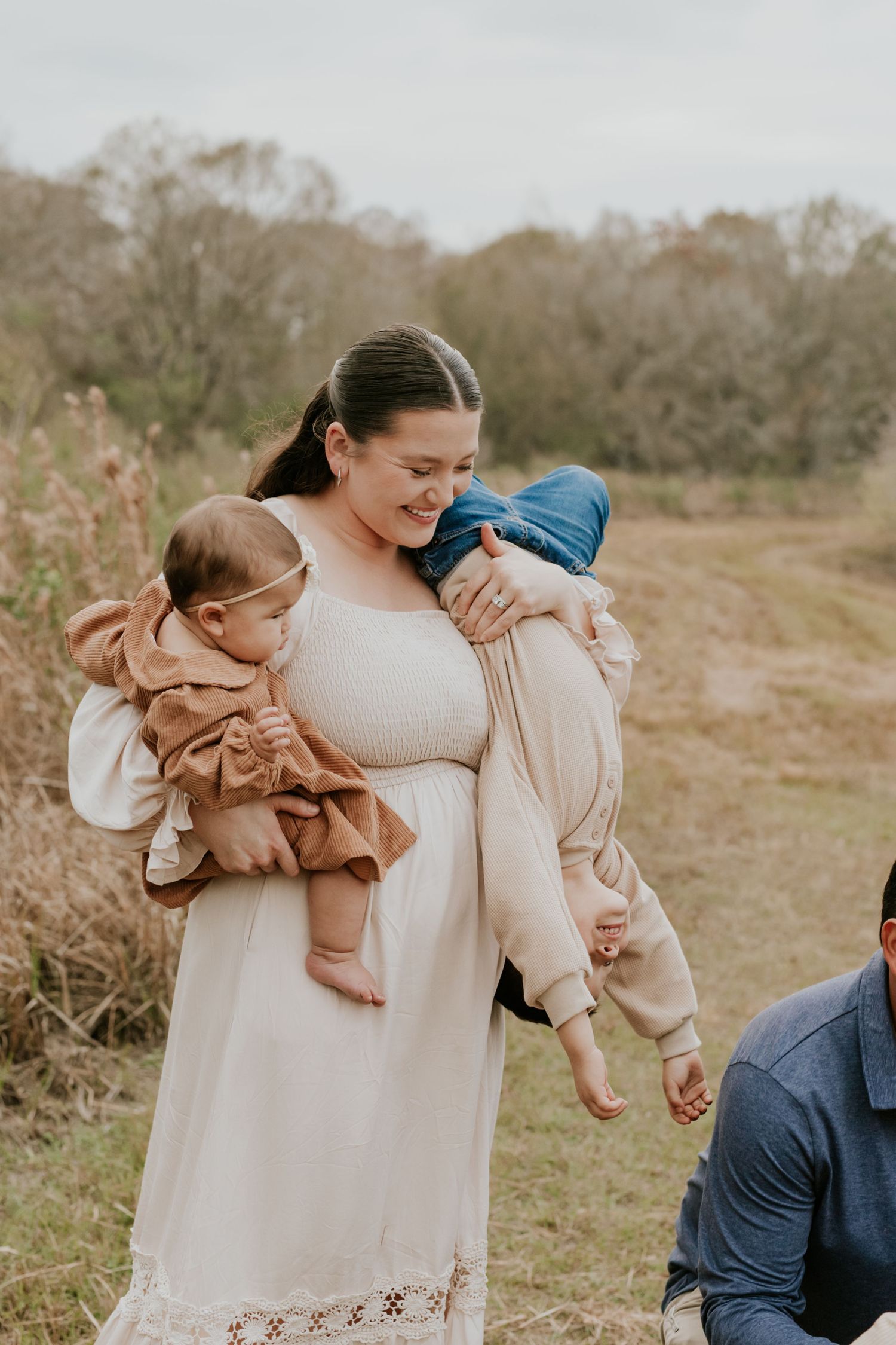 Mother in white dress carrying baby and toddler in autumn field setting.