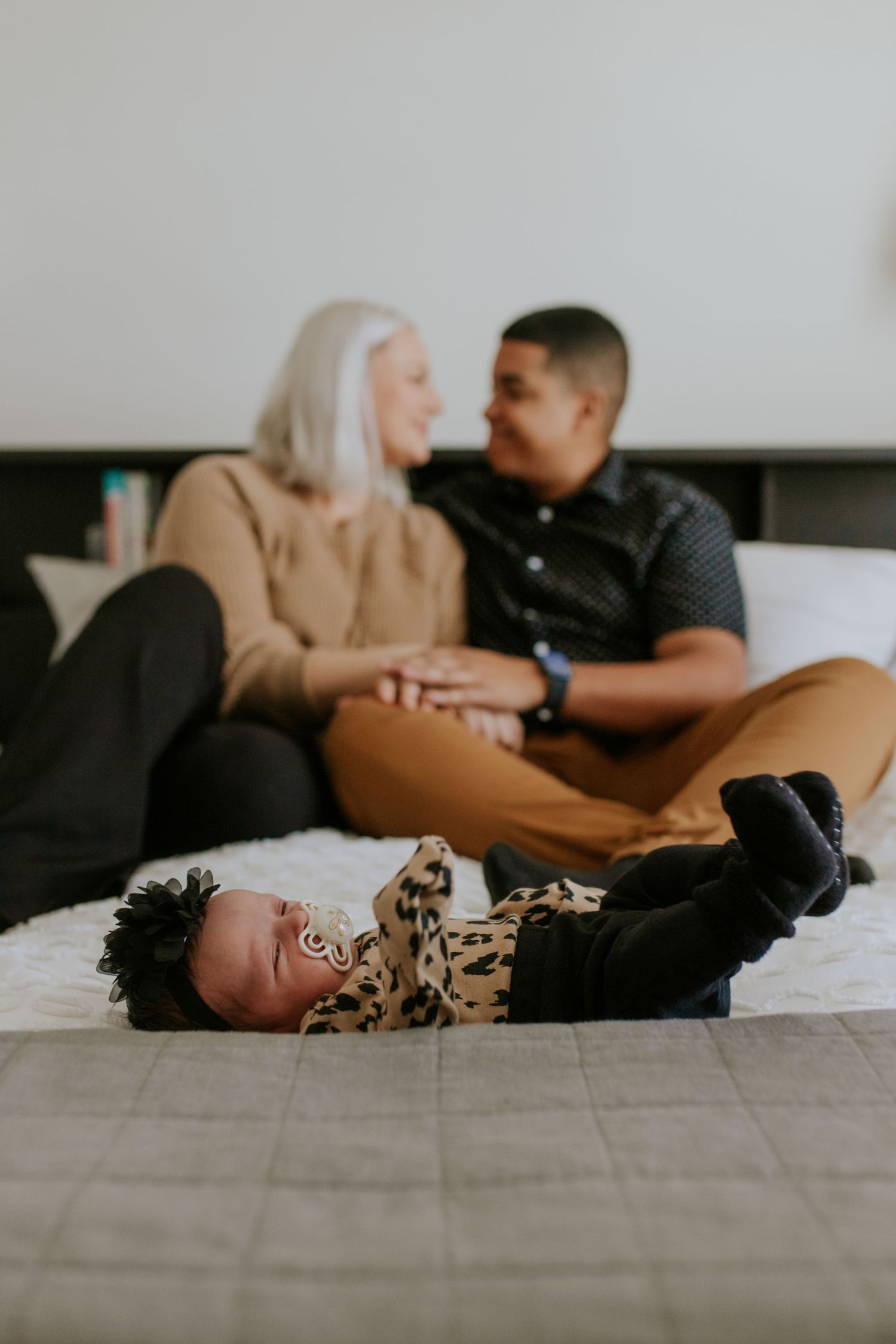 Newborn in leopard outfit lies on bed as parents share a loving glance behind them.
