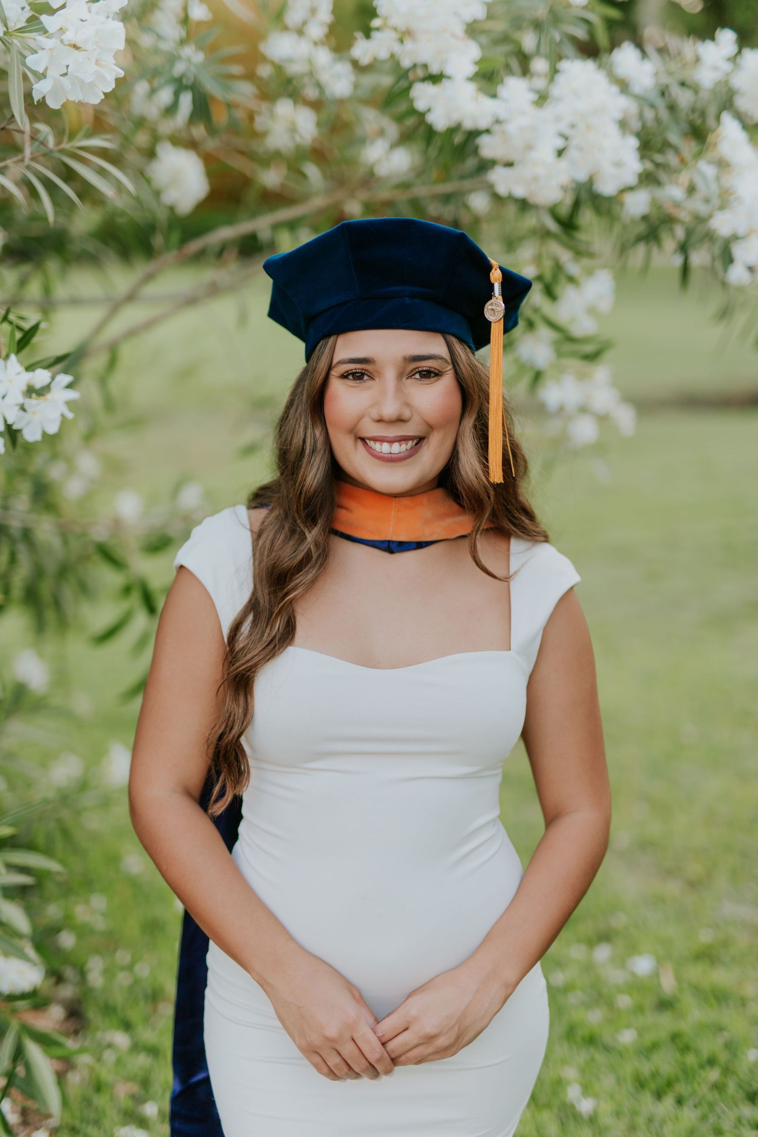 Smiling graduate in white dress and navy cap with gold tassel poses outdoors among white flowering trees.