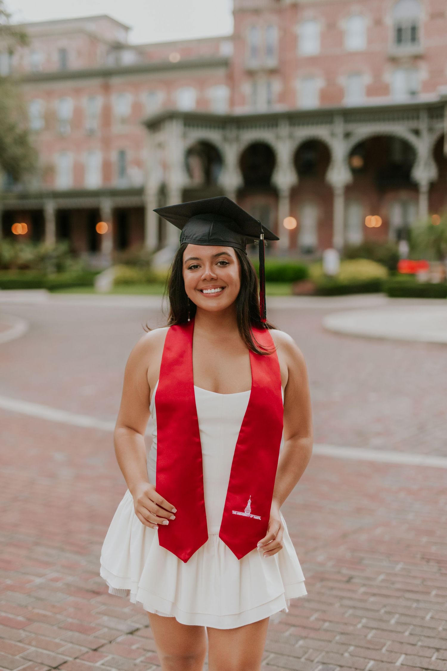 Smiling graduate in white dress, black cap, and red stole poses in front of historic building.