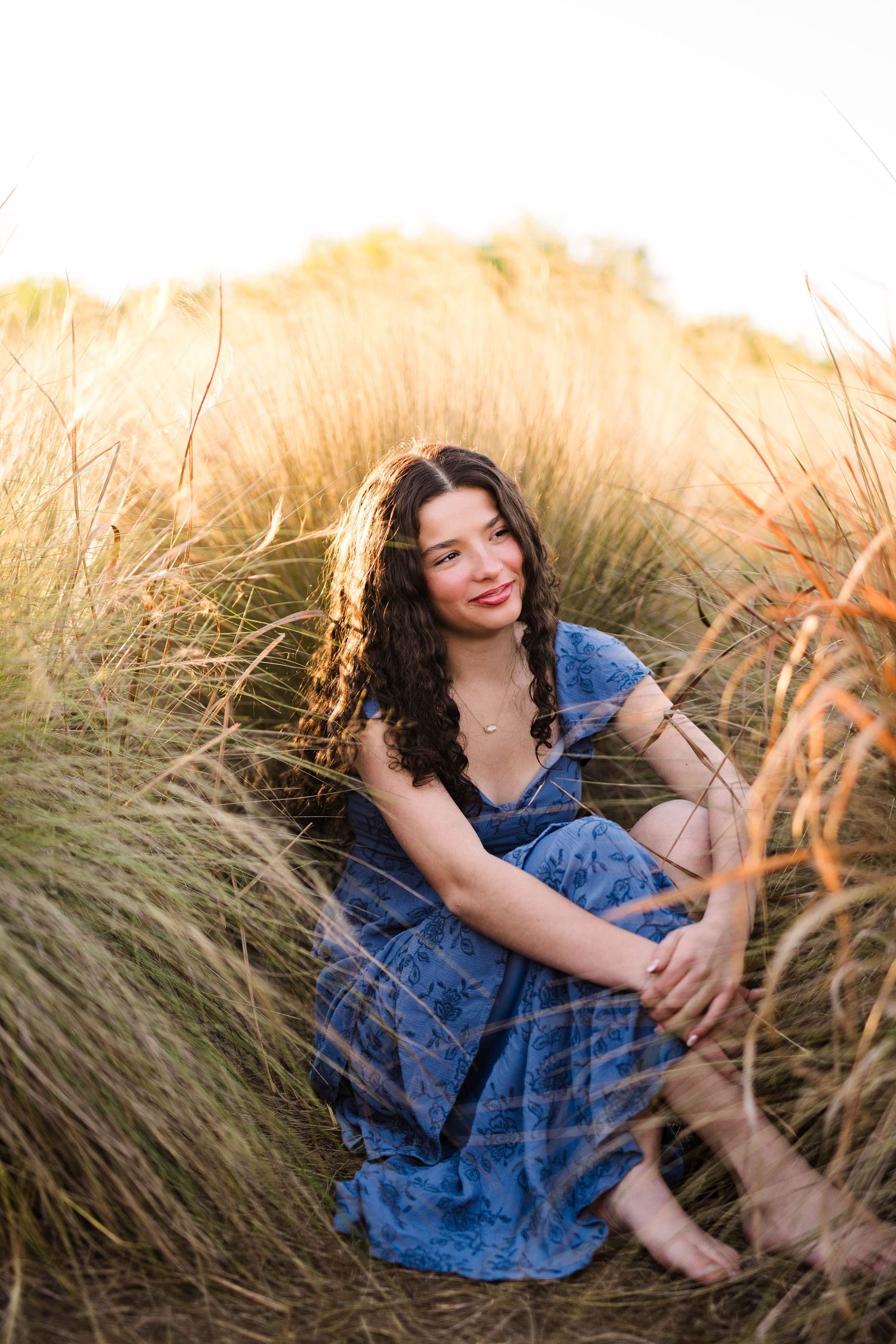 Young woman in blue floral dress sitting barefoot in golden tall grass at sunset smiling.