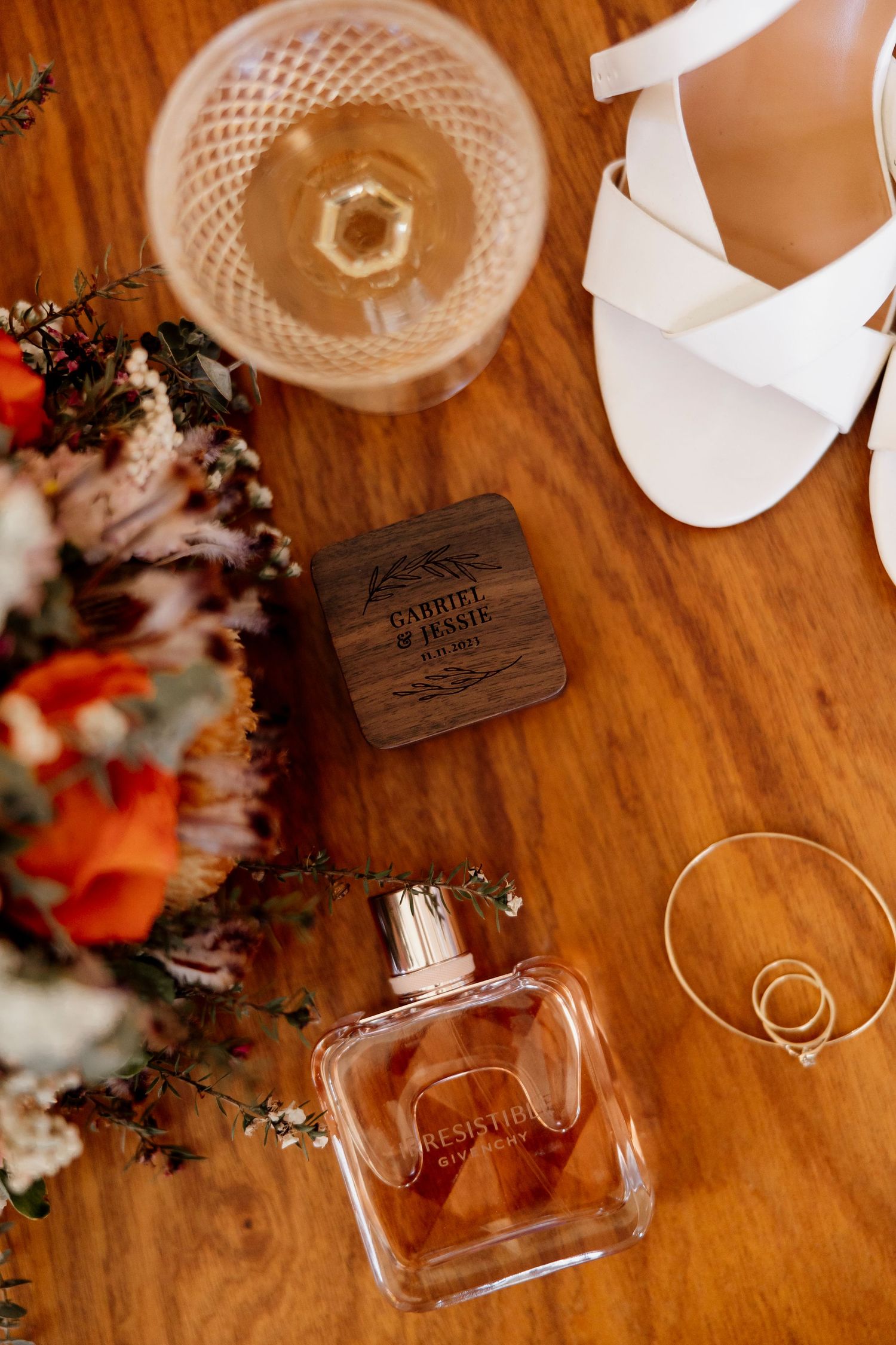 Bridal details flatlay with perfume, gold earrings, white heels, champagne and a personalised wooden ring box — Oh Joy! Photography Tauranga